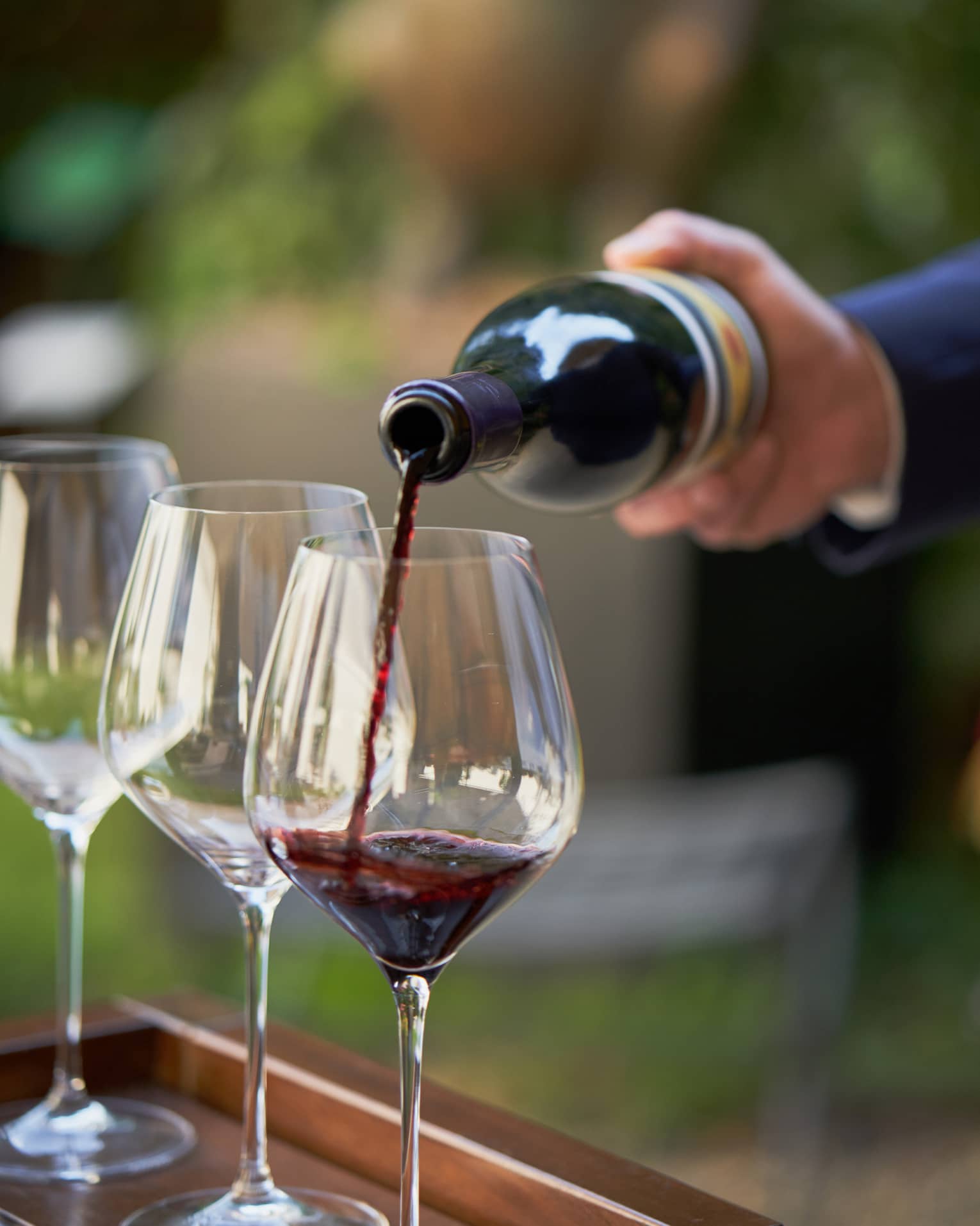 A close up of hands pouring a bottle of red wine into a clear wine glass that rests on a wooden tray, outdoors