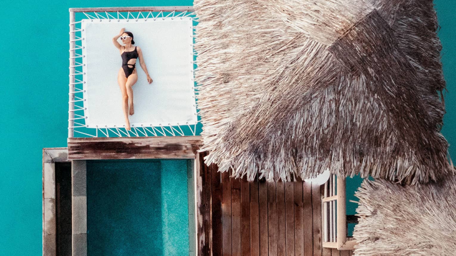 Woman lays on overwater net next to thatched-roof bungalow