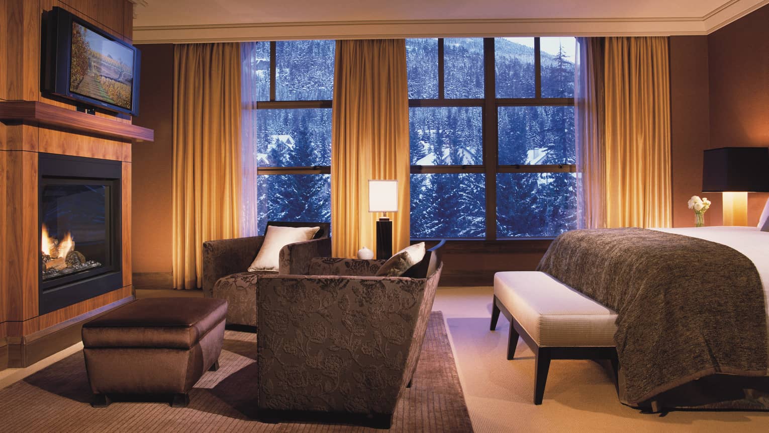 Edge of hotel room bed, sofa and ottoman facing TV and gas fireplace, snowy mountain views from window