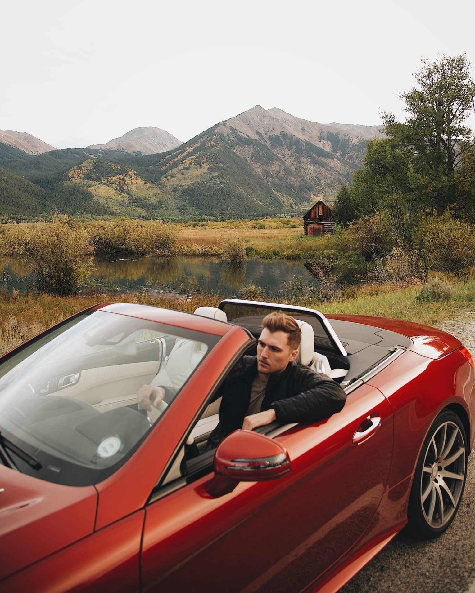 Driver in a shiny, cherry red two-door convertible with the top down, a marshy valley and forested peaks in the distance.