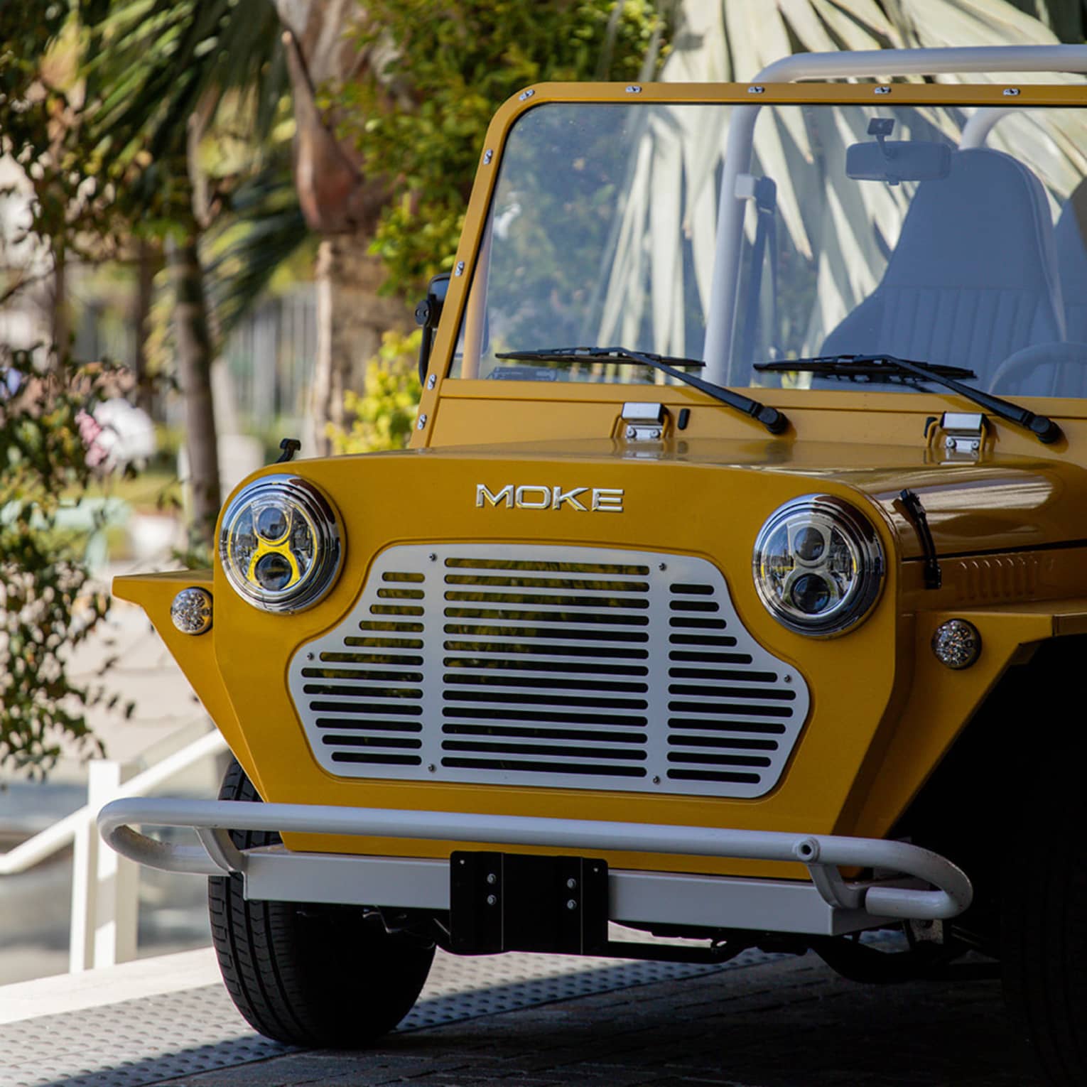 Extreme close-up of a dark-yellow Moke parked in the shade against a backdrop of palm trees and other greenery.