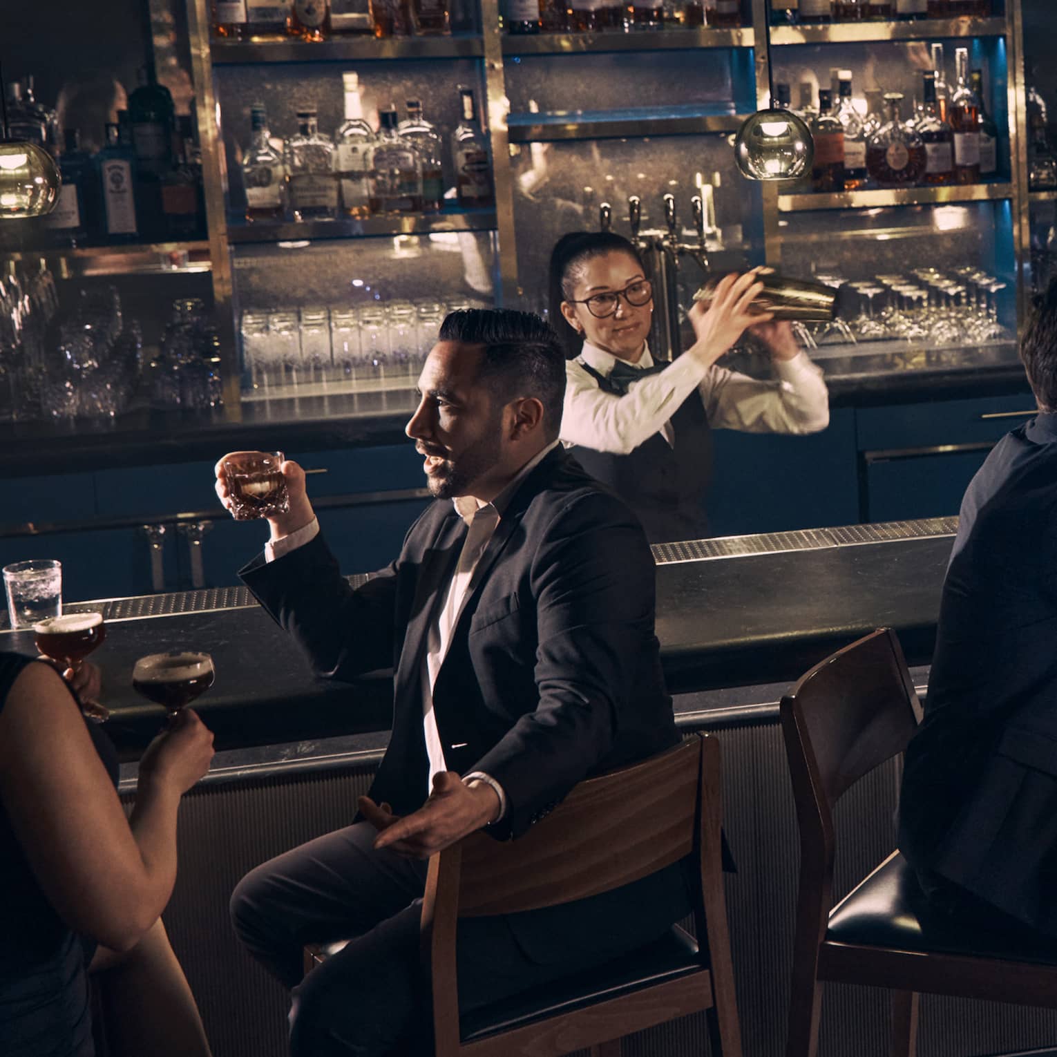 Guests chat over drinks at the bar at Prosperous Penny as a bartender shakes a cocktail shaker behind the bar.