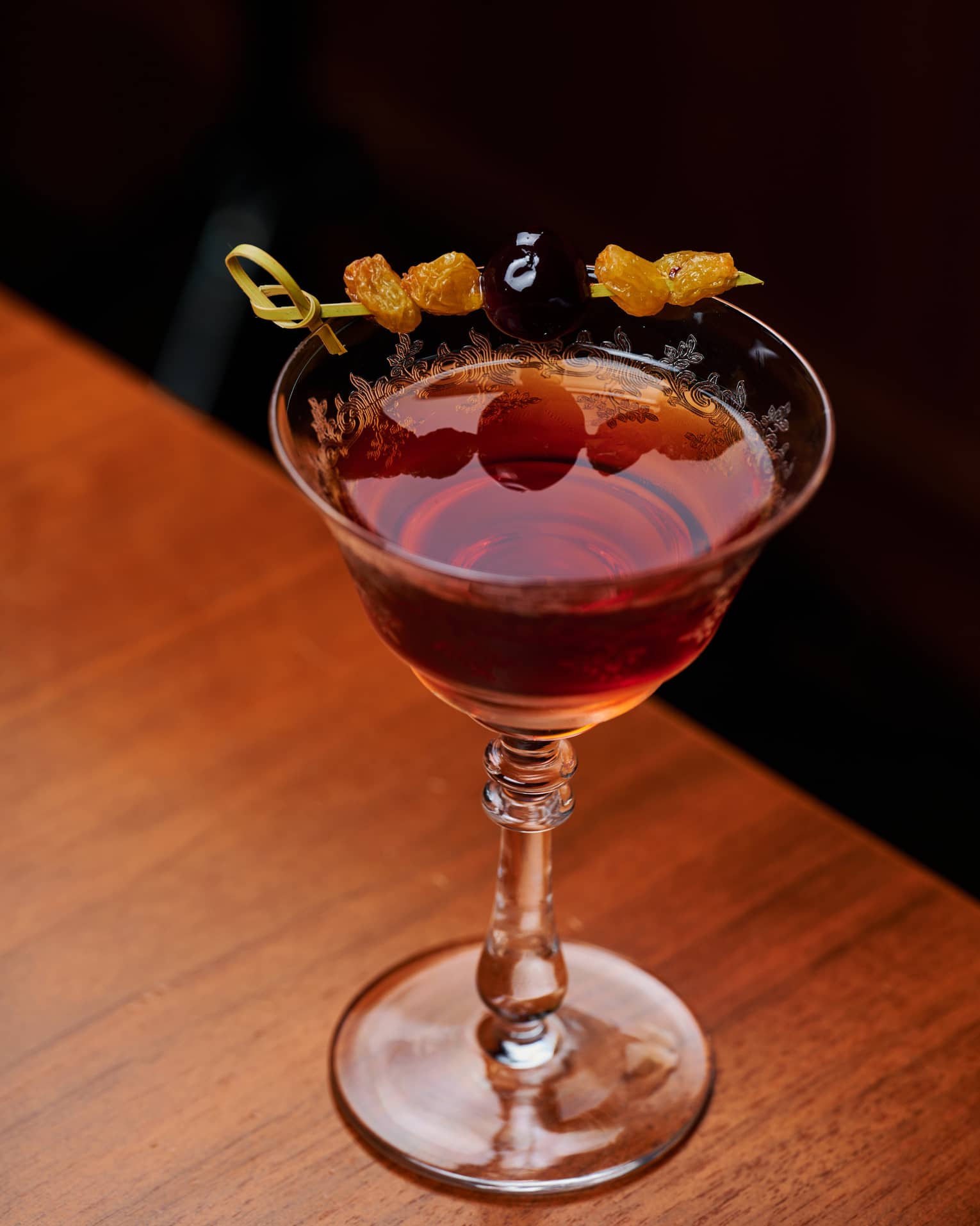Close-up of a red cocktail in a finely engraved stemmed glass garnished with a skewer of raisins and a brandy cherry.