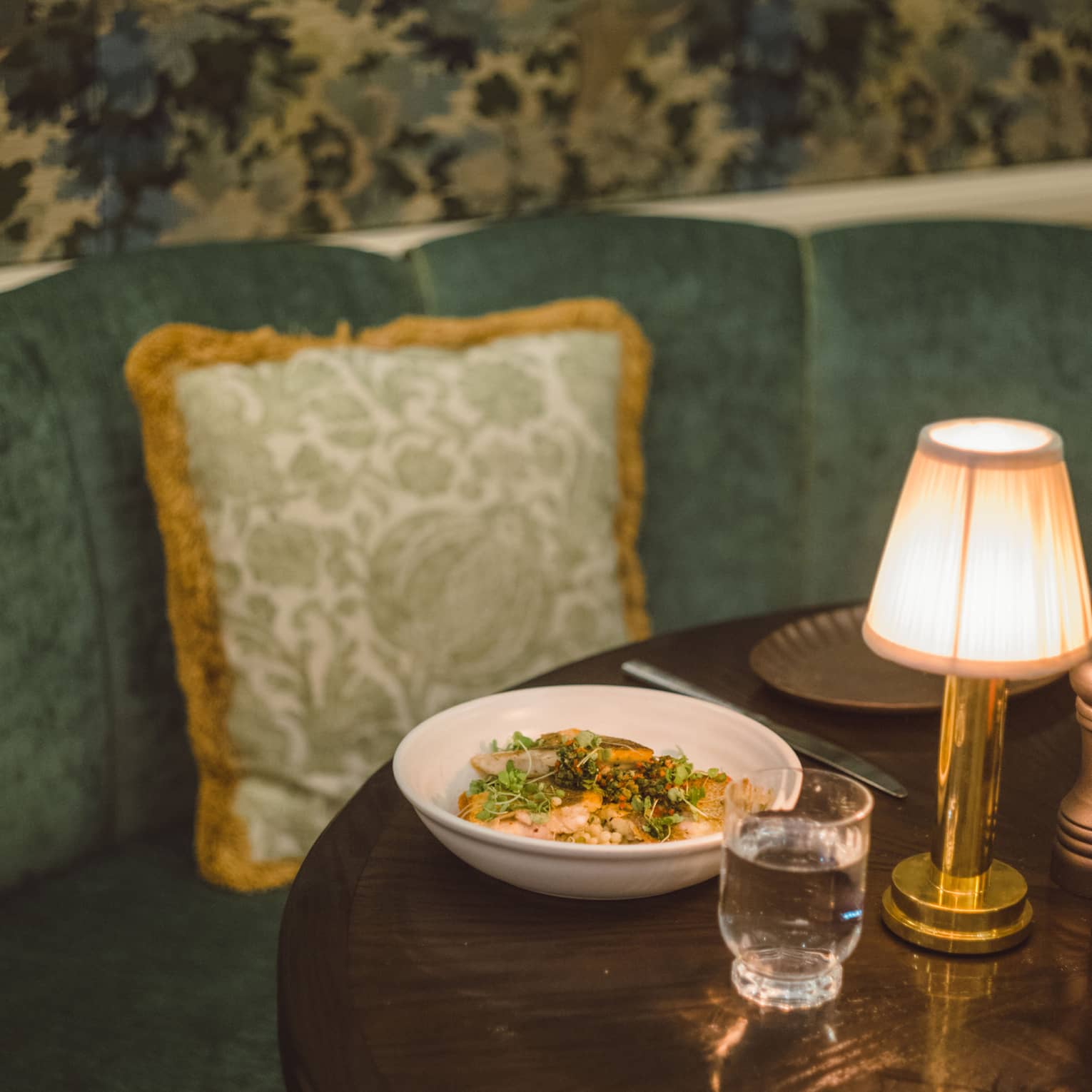 A colourful dish sits atop a wooden, lamplit table with a green velvet booth behind