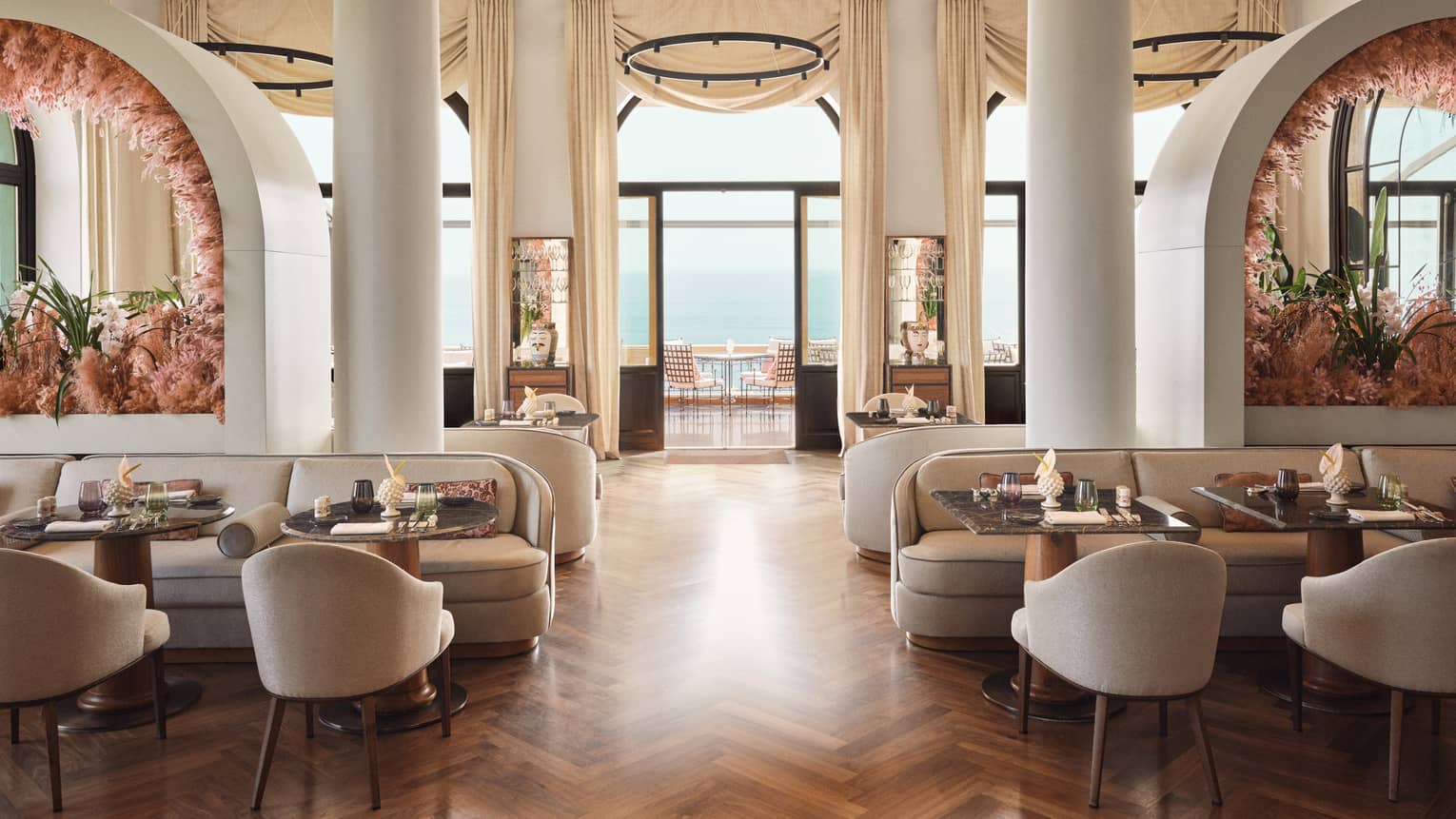 Restaurant dining room with two large columns in the centre, parquet wooden flooring, tall ceilings and a wall of windows that opens up to a terrace overlooking the sea