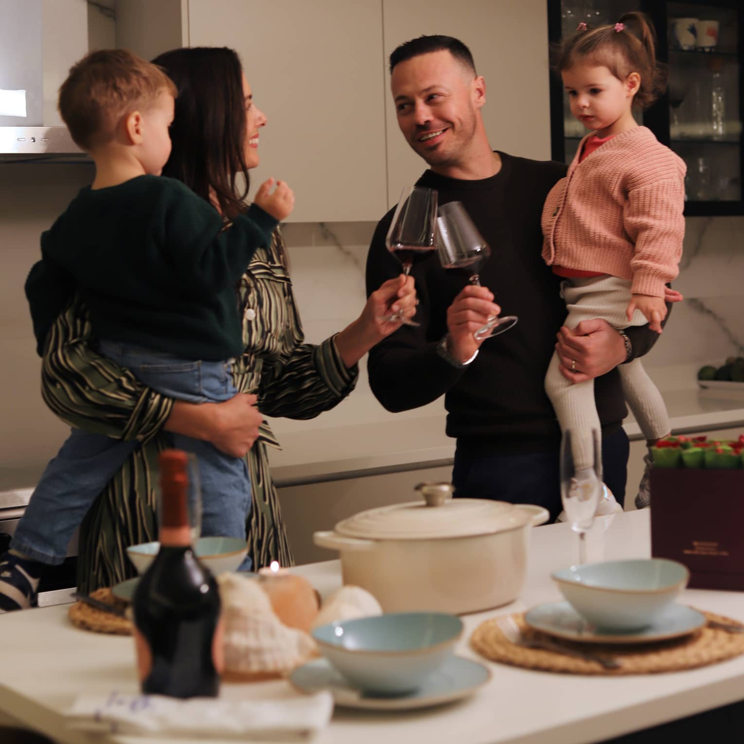 Two adults, each holding a small child in their arms, smile at each other and click their glasses of wine together while standing behind a kitchen counter