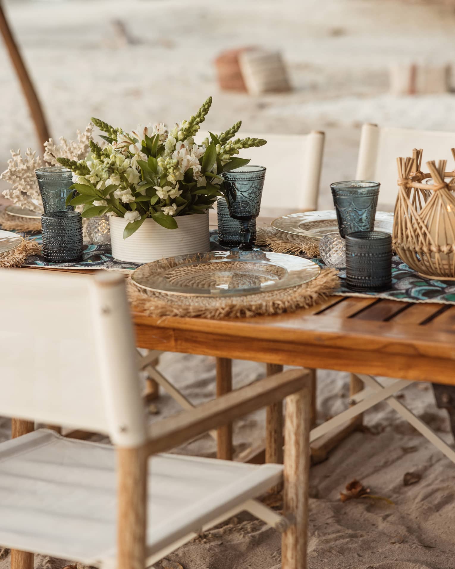 Dining table on private beach set with teal-blue vintage goblets, clear glass plates on woven mats and a bouquet of flowers.