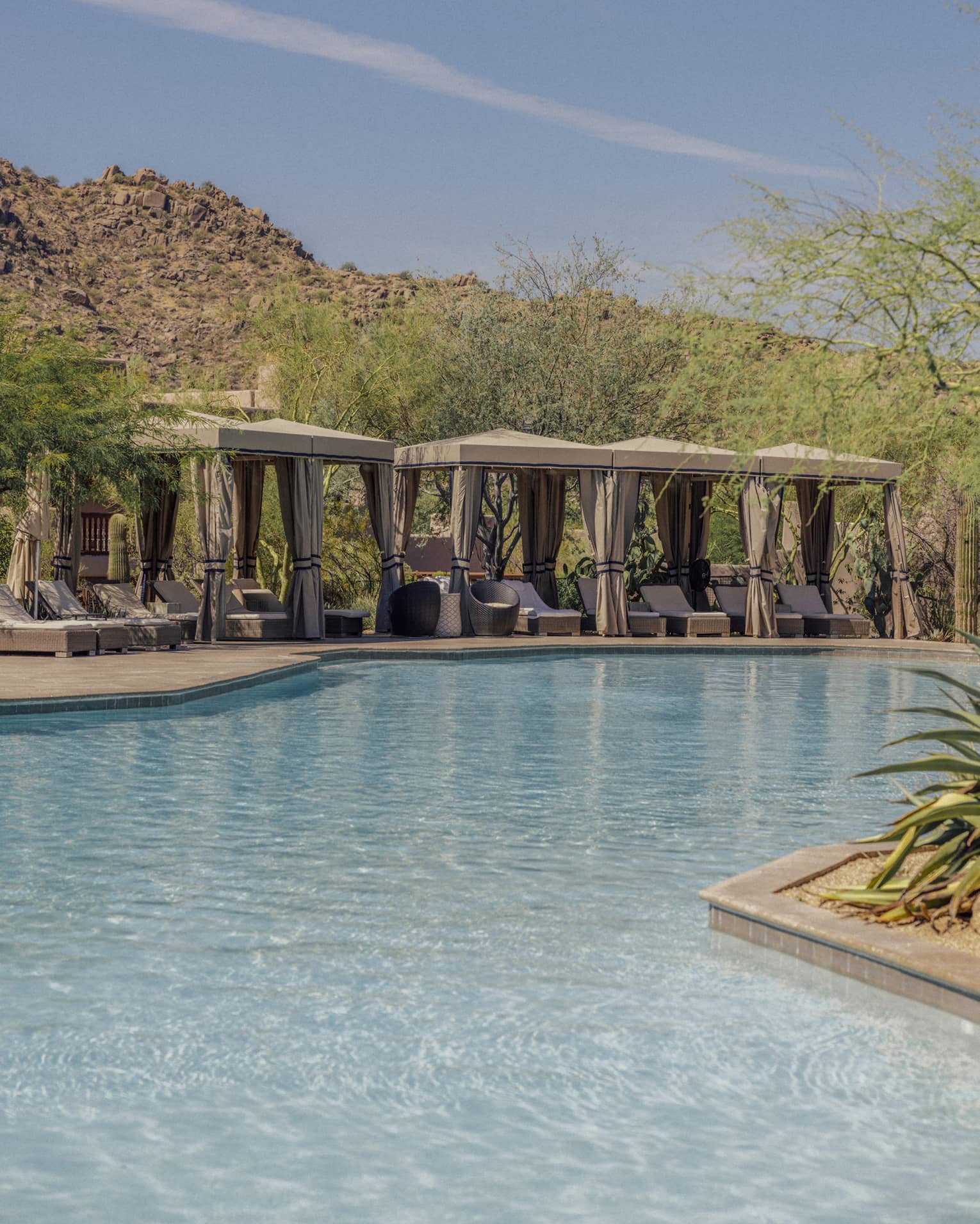Four cabanas with lounge chairs near the pool with mountains in the background.
