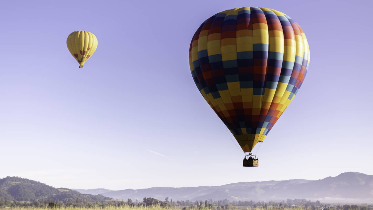 Close-up of a multi-coloured hot-air balloon close to the ground; a second yellow and red balloon floats higher above it.