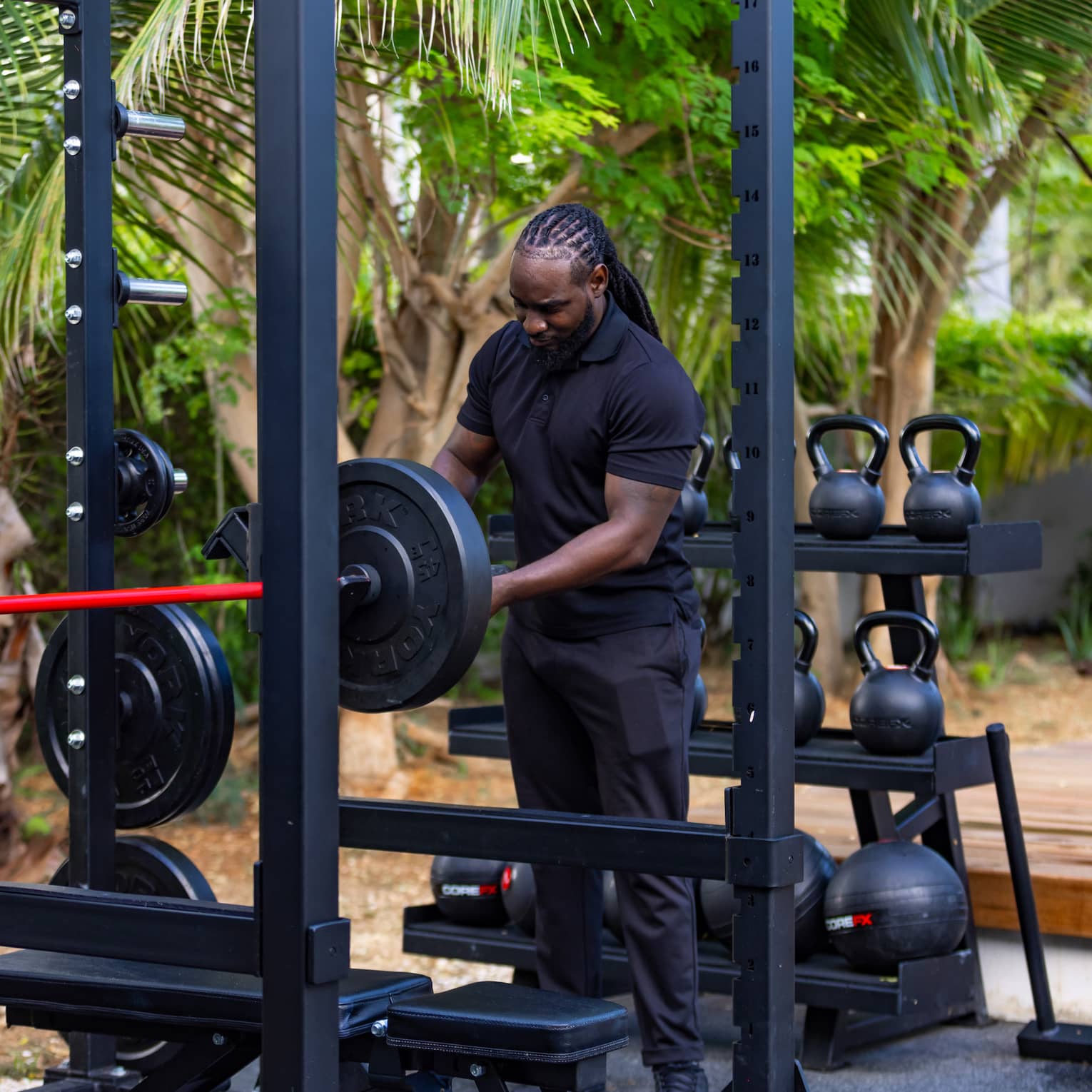A guest adjusts a barbell in an outdoor gym against a rack of kettlebells and medicine balls and a grove of shady trees.