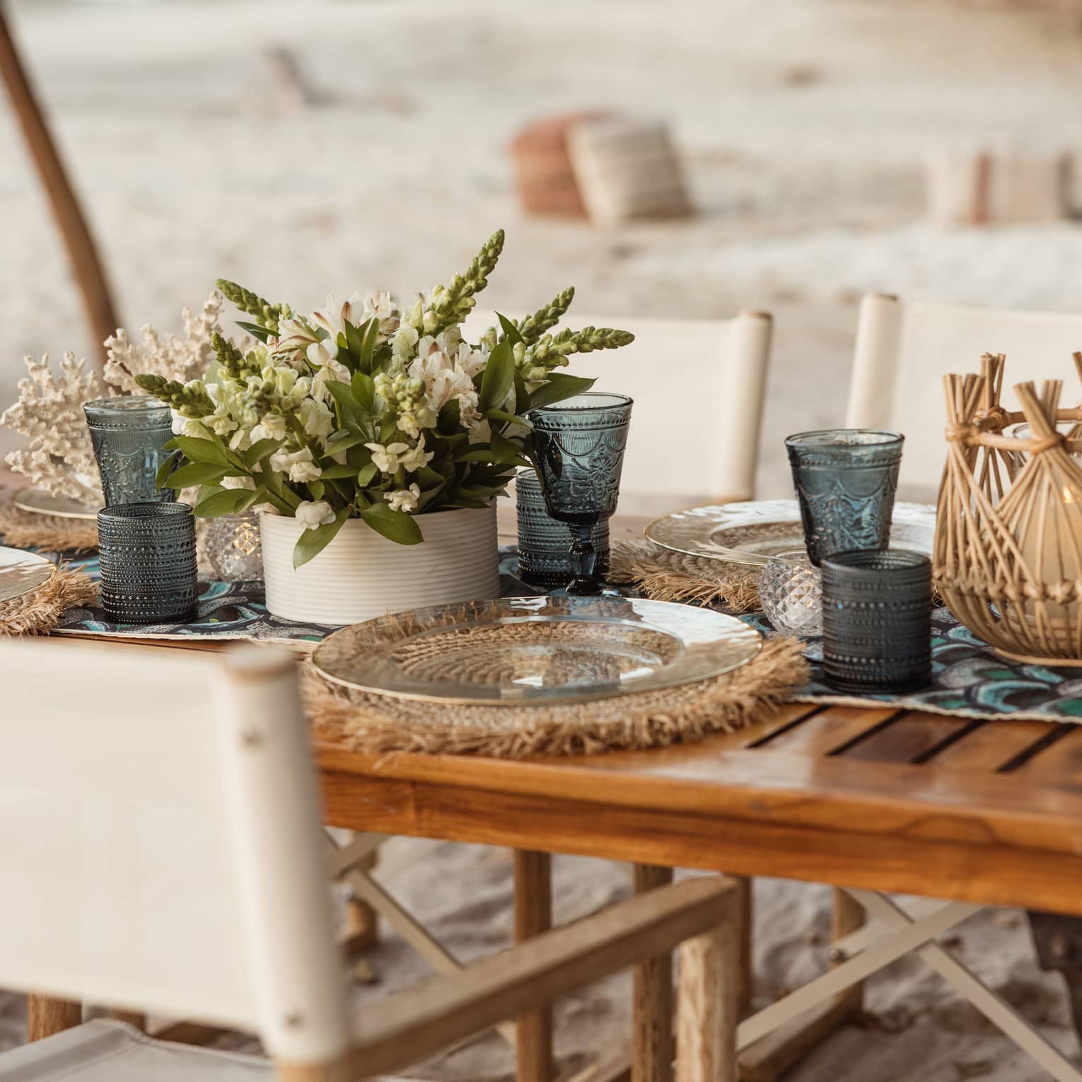 Dining table on private beach set with teal-blue vintage goblets, clear glass plates on woven mats and a bouquet of flowers.
