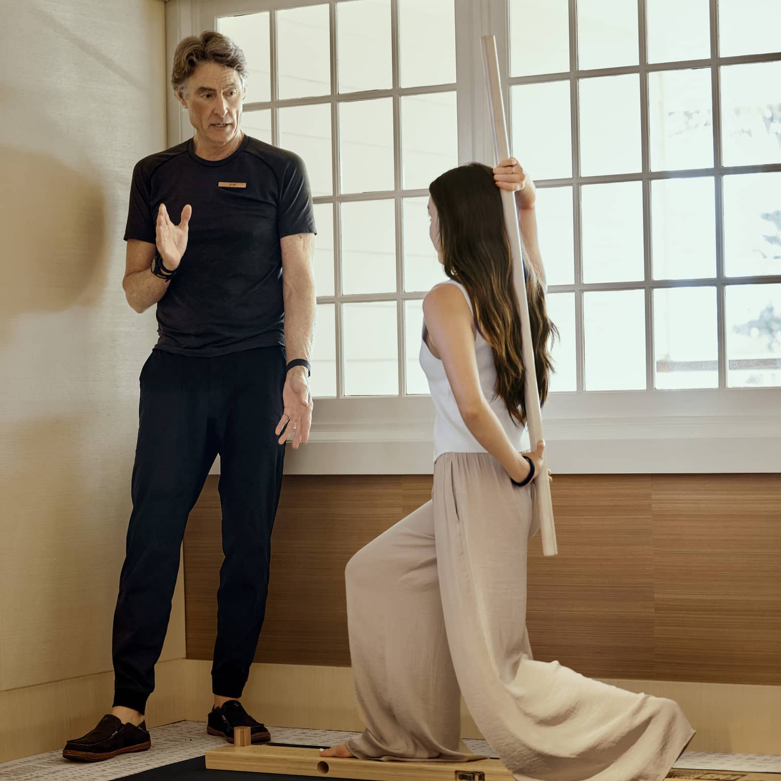 Facing an instructor, a guest stands in a high lunge on a narrow wood plank, gripping a dowel vertically behind their back. 