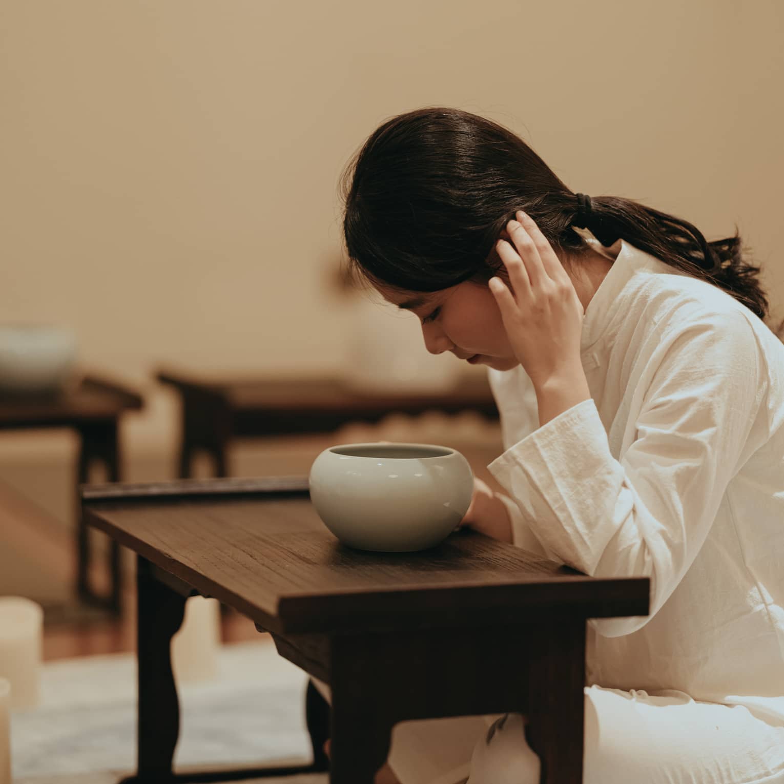 Cross-legged at a low wooden table, a barefoot practitioner in white leans in and cups an ear over a rounded ceramic bowl.