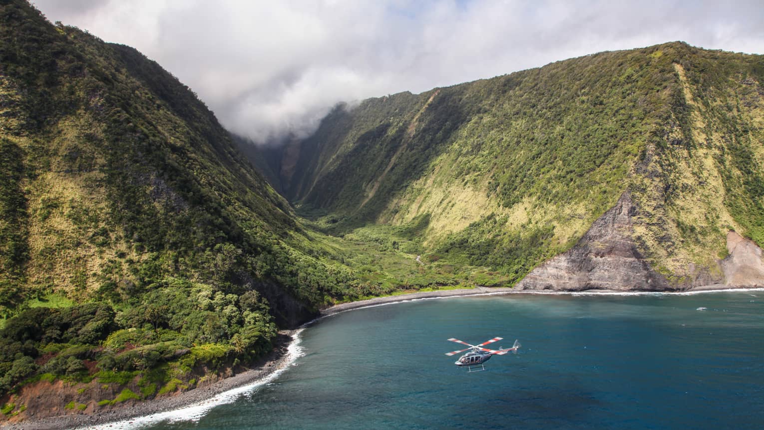 Helicopter flies over ocean by sweeping green volcanic mountains