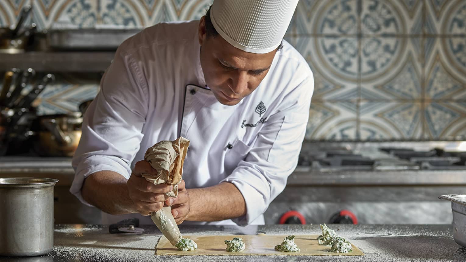 Chef squeezes batter from piping tube onto tray in kitchen with patterned tile wall