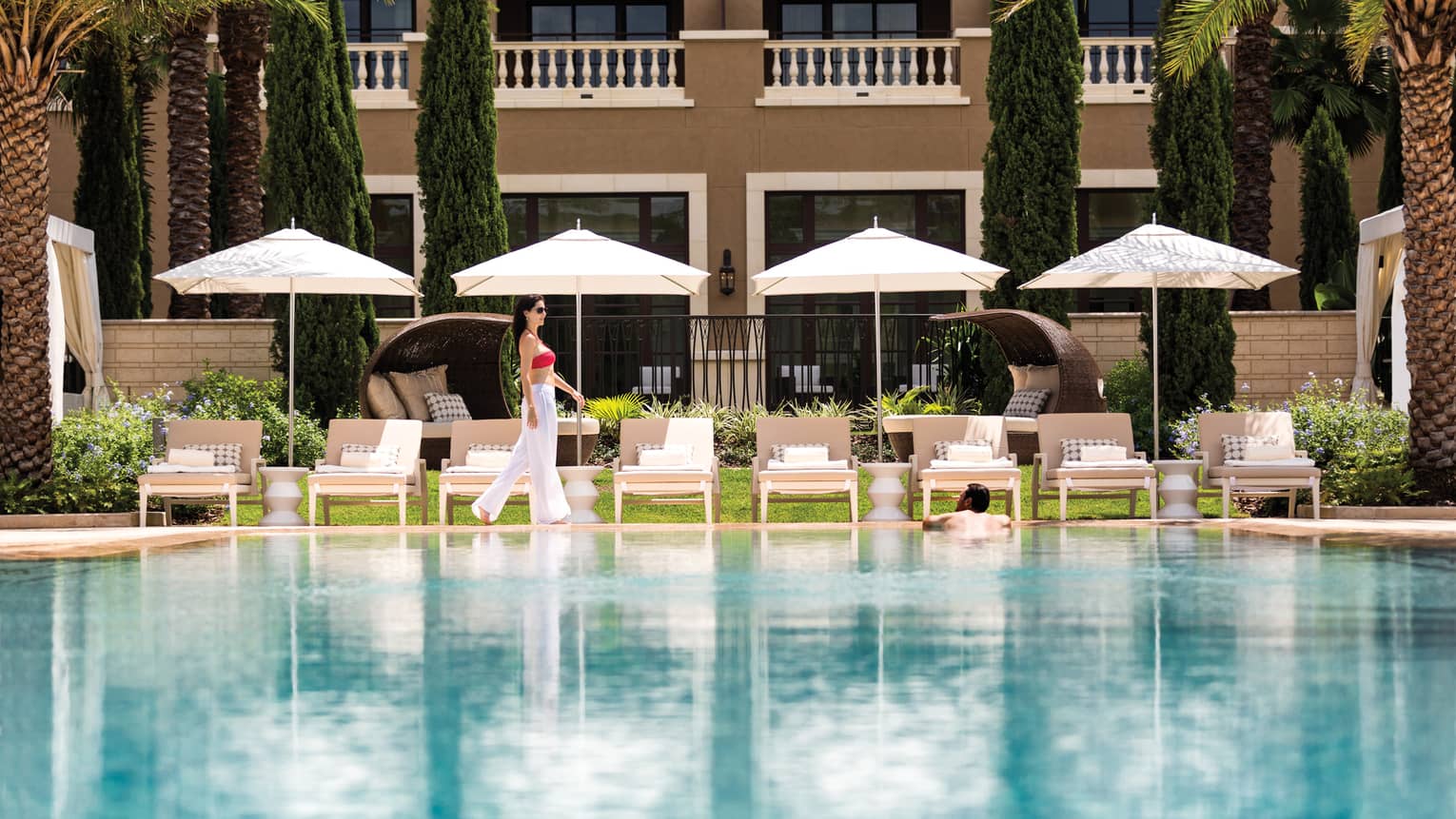 Woman walks by row of white patio chairs, umbrellas on outdoor swimming pool deck