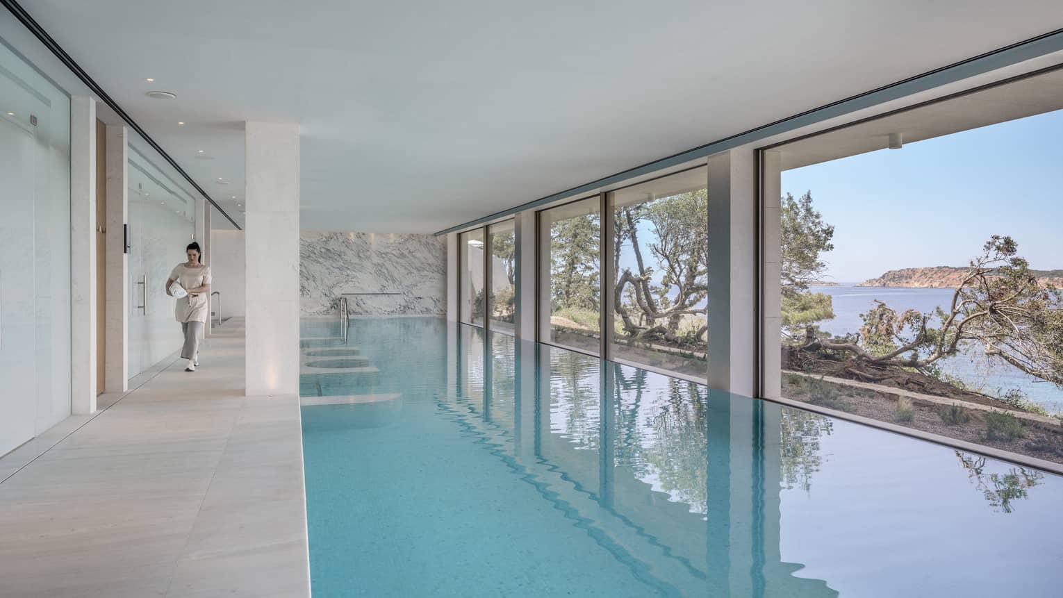 A guest walks on the left side of a spacious indoor pool. A wall of windows on the right reveals twisted trees and the ocean.