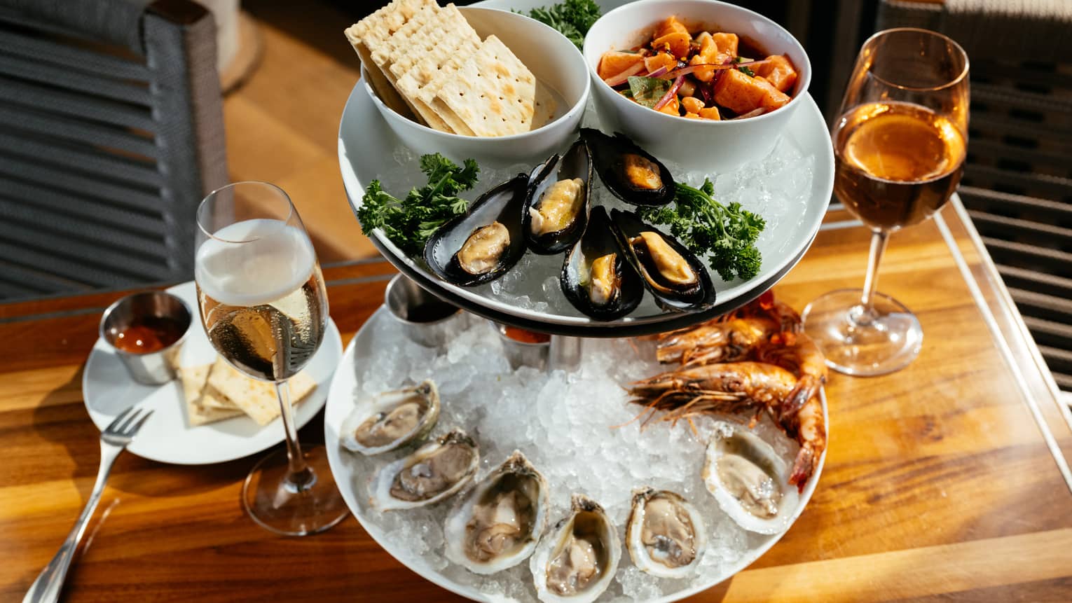 A two-tiered serving tray with assorted seafood items, crackers and garnishes. Two stemmed glasses with drinks and a small plate for crackers and sauce are next to the tray.