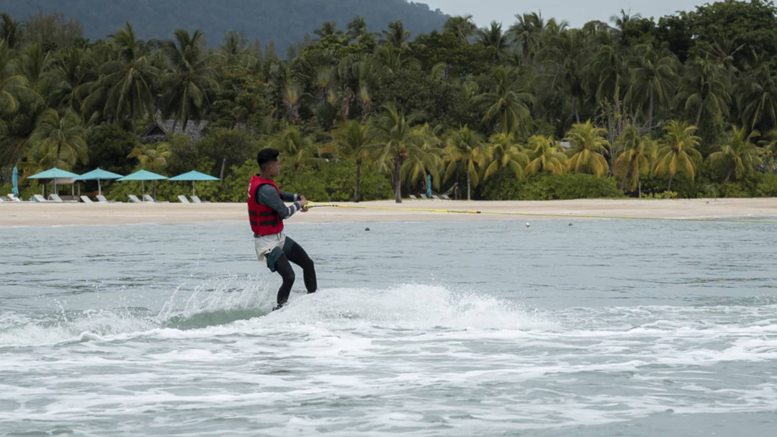 Person in red life jacket glides along the water on a water ski, the shore and copse of palm trees in the distance