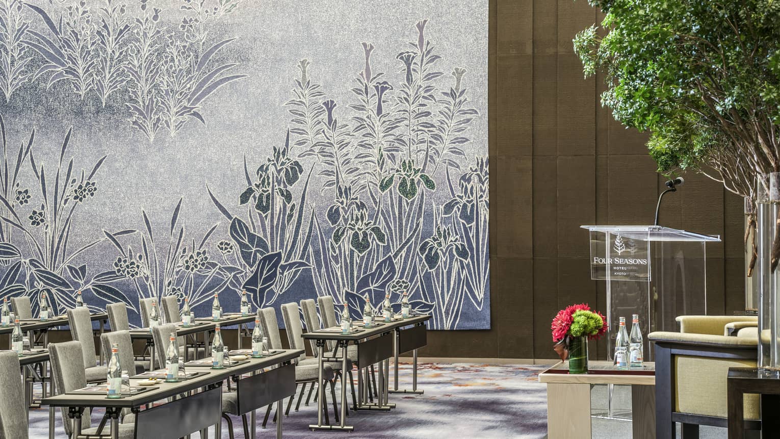Conference in ballroom, rows of meeting tables, chairs along floral accent wall, facing podium