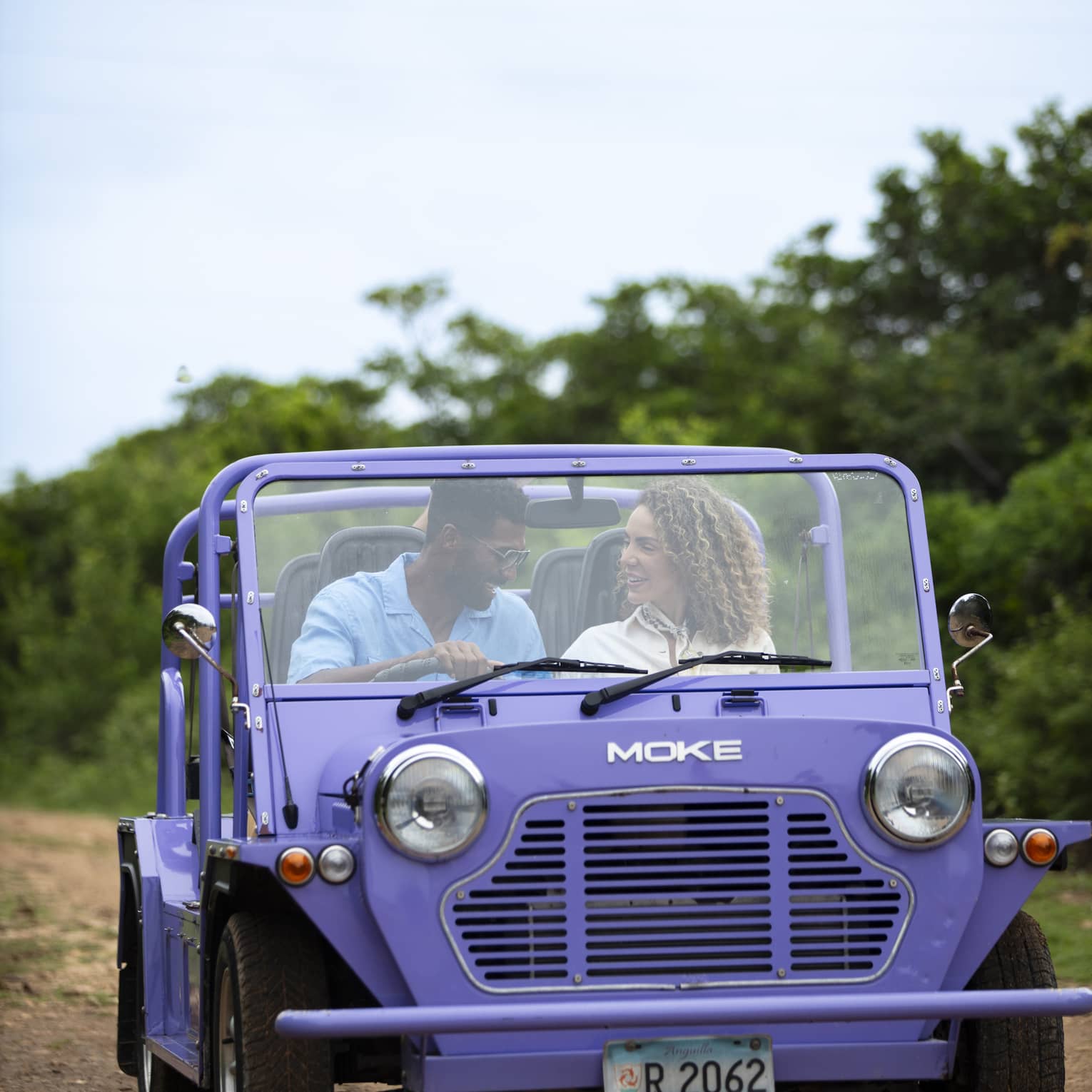 Front view of a couple driving a purple Moke, a small open-air recreational vehicle, along a dirt road surrounded by trees.