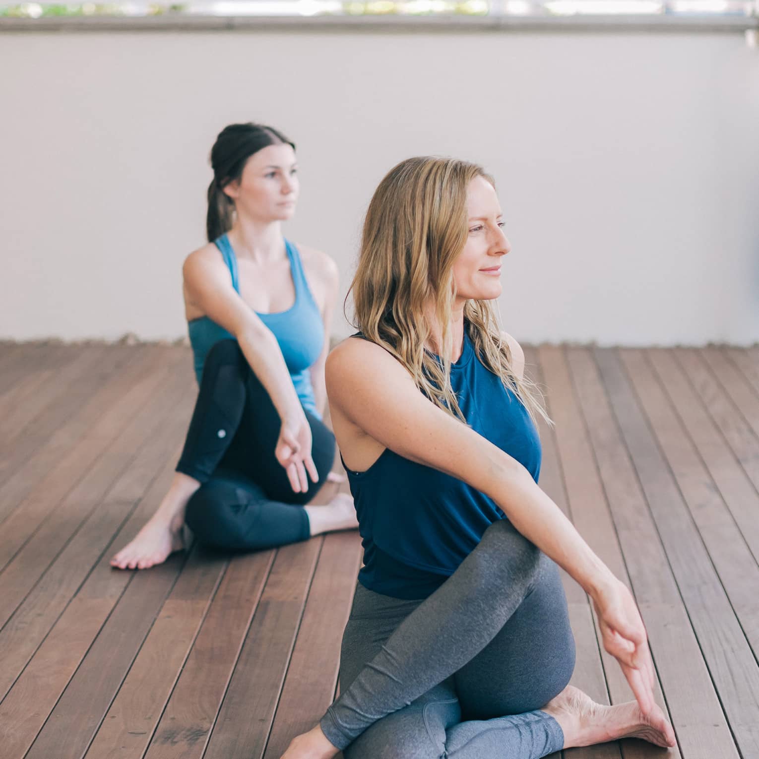 One blonde woman and one brunette woman sit with one leg crossed over the other with their bodies turned to one side and their front hands forming a lam mudra