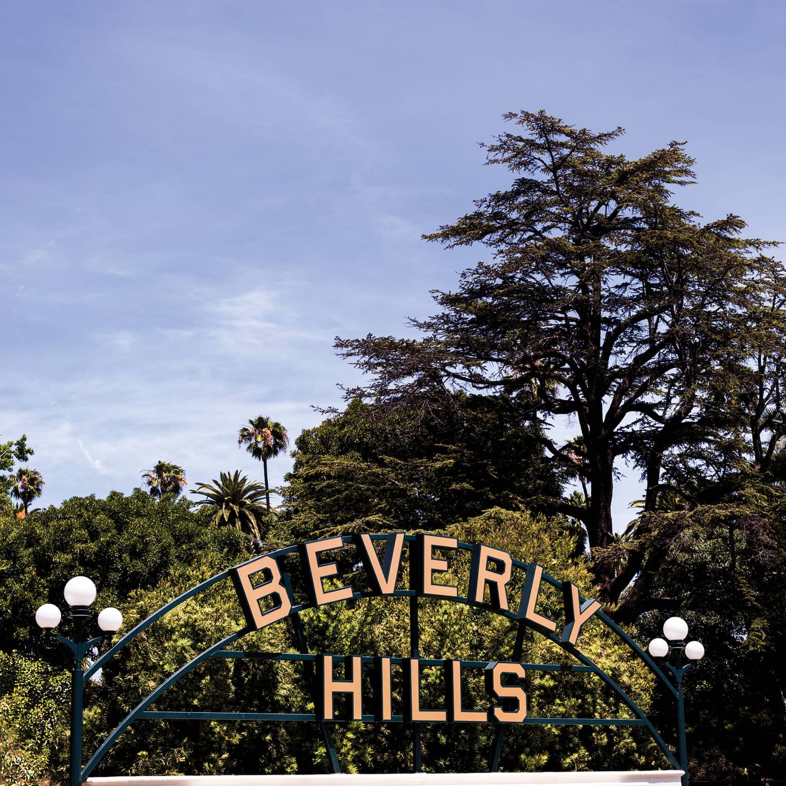 Large pink letters, mounted on a metal frame, spell out the name "Beverly Hills" against an outdoor backdrop of leafy trees.
