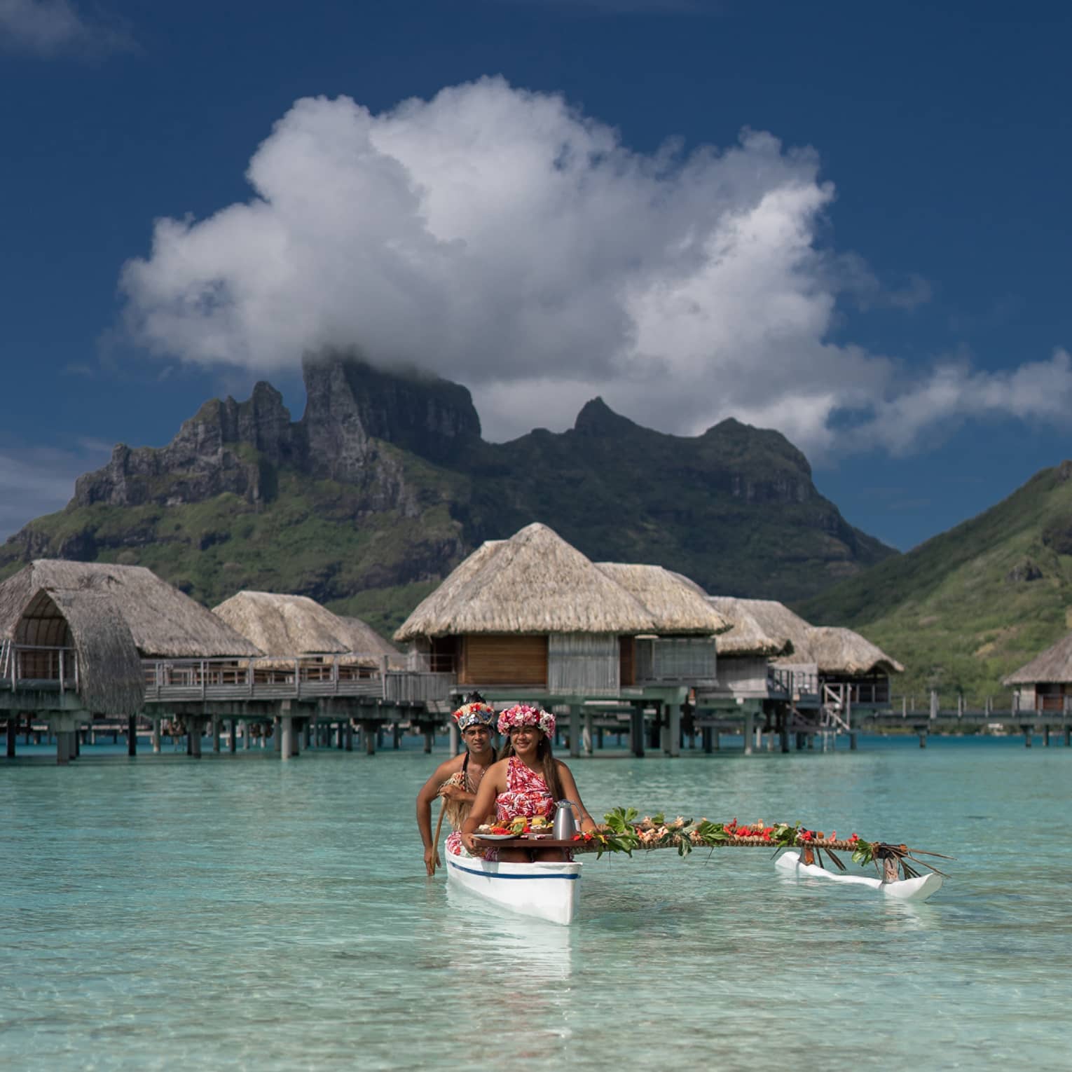 Two vibrantly dressed people in a canoe proffer a tray with colourful food attached to a pole draped with leaves and flowers.