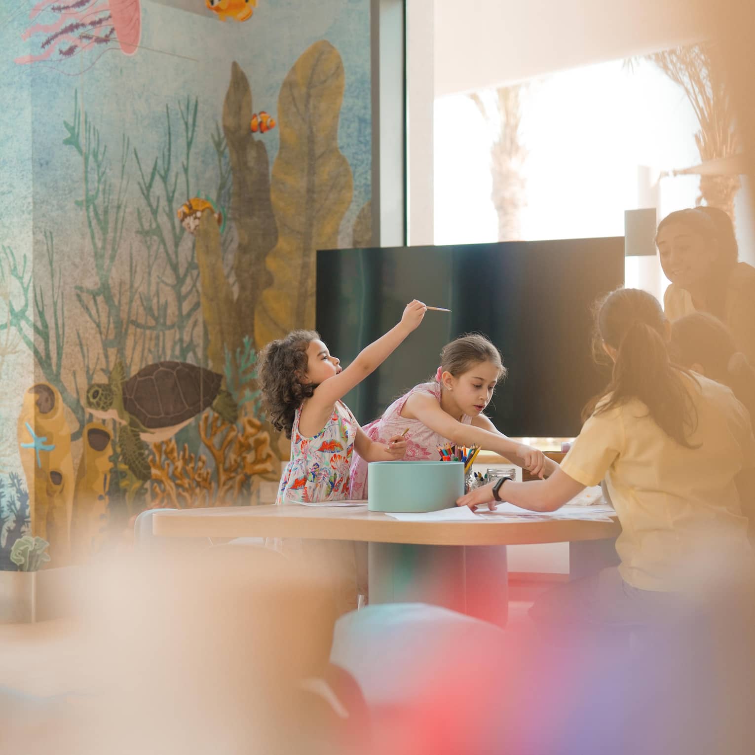 Two young children sit at a crafts table while an adult helps them paint