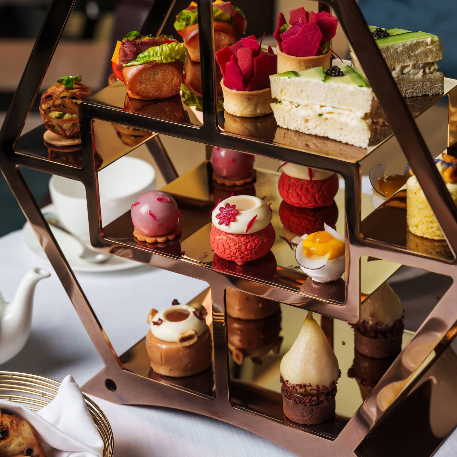 Bronze hexagonal three-tiered tray filled with small pastries and set on a table next to small containers of jam and a silver bread basket
