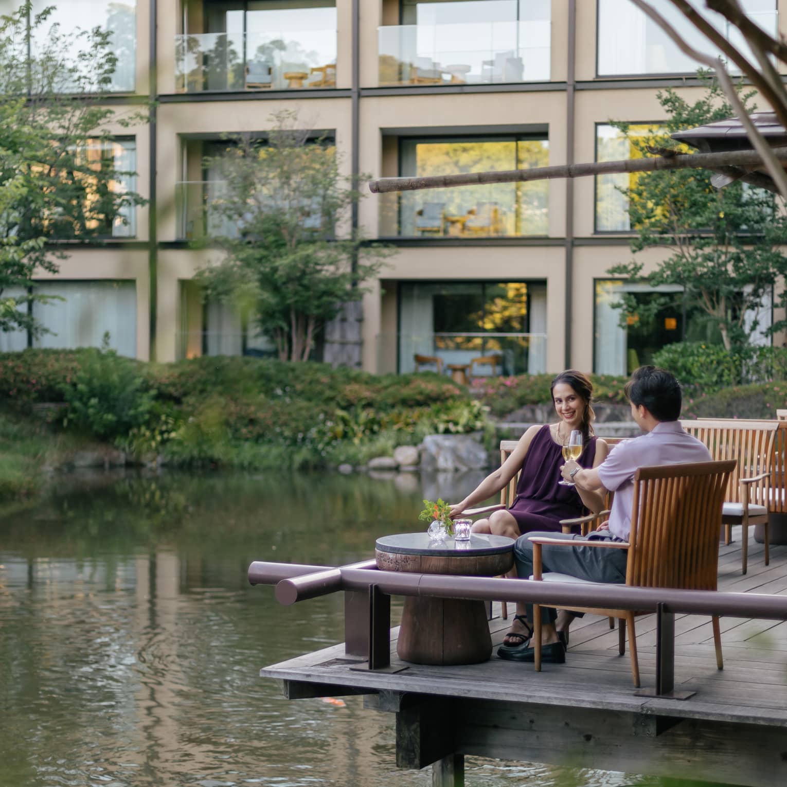 Smiling couple clink champagne flutes at a café table on a wood deck overlooking a rippling pond bordered by lush foliage.