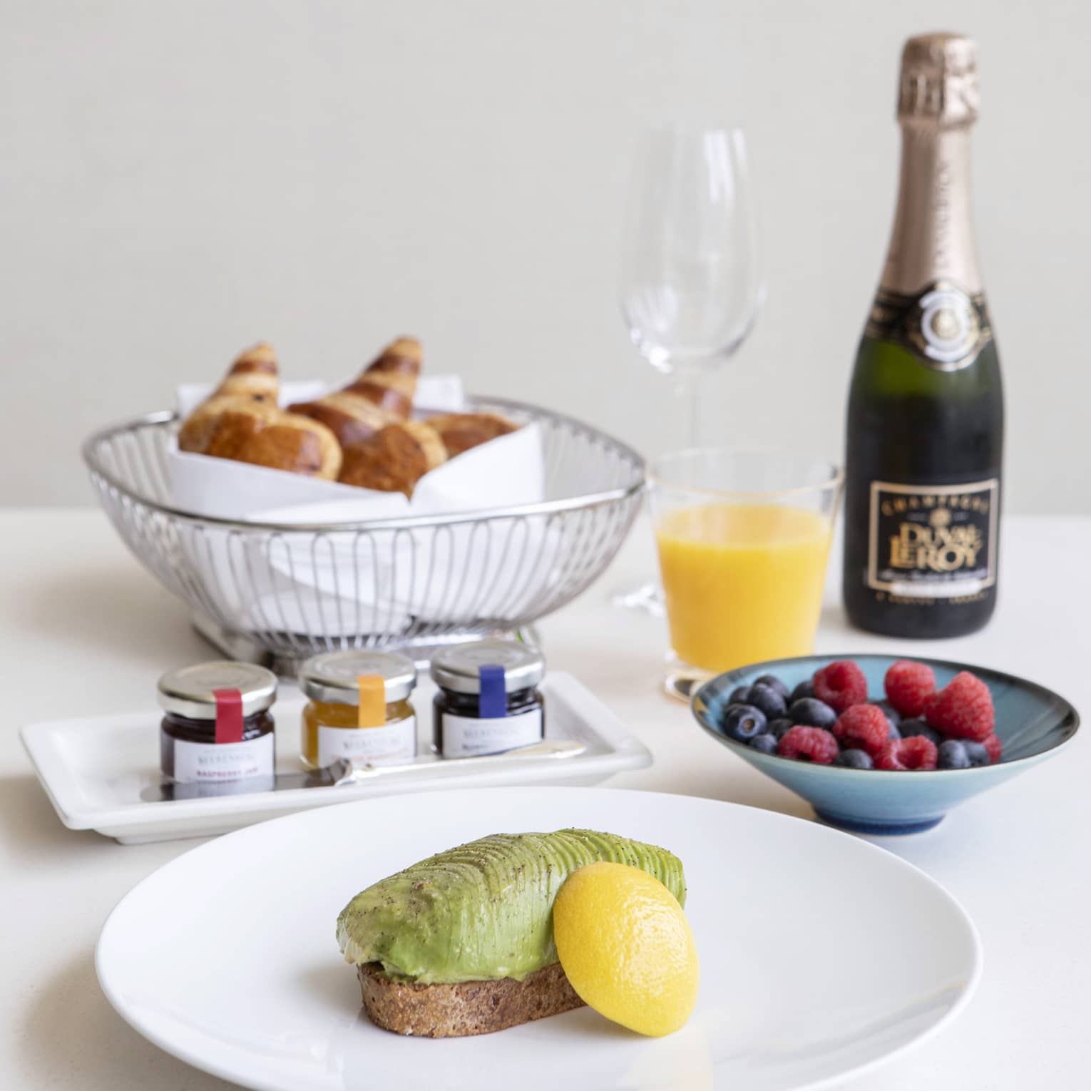 Luxurious breakfast including avocado toast, a basket of croissants, fresh fruit, and champagne and orange juice.