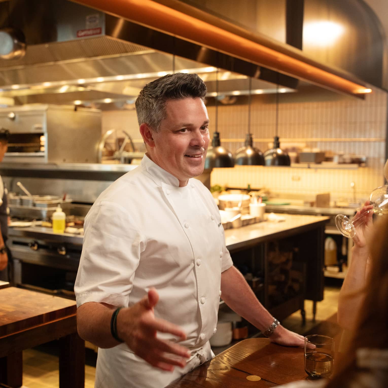 Chef standing beside a dining table in the kitchen talking to guests.