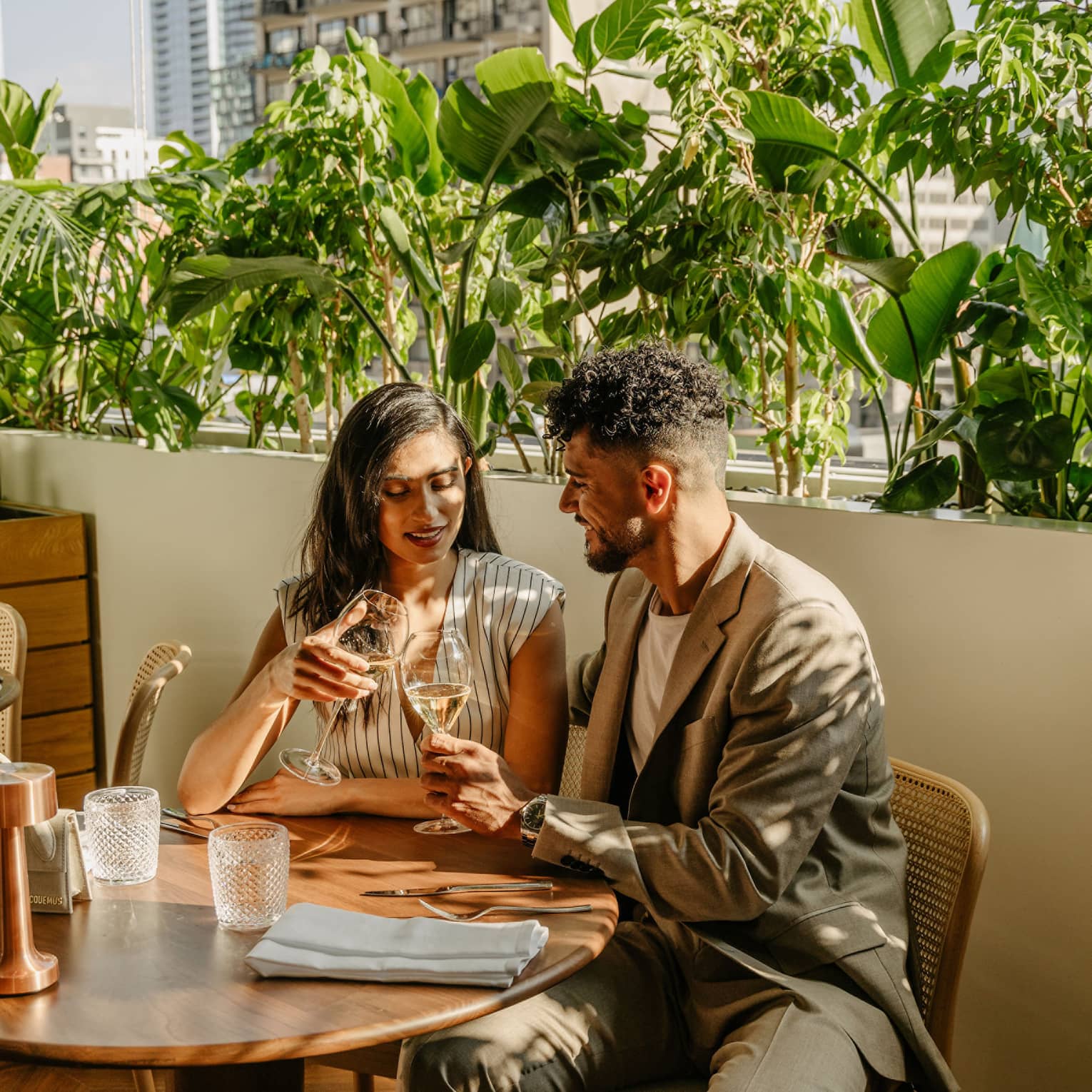 A smiling couple clink glasses of white wine as they sit side by side on an sun-dappled outdoor patio bordered by foliage.