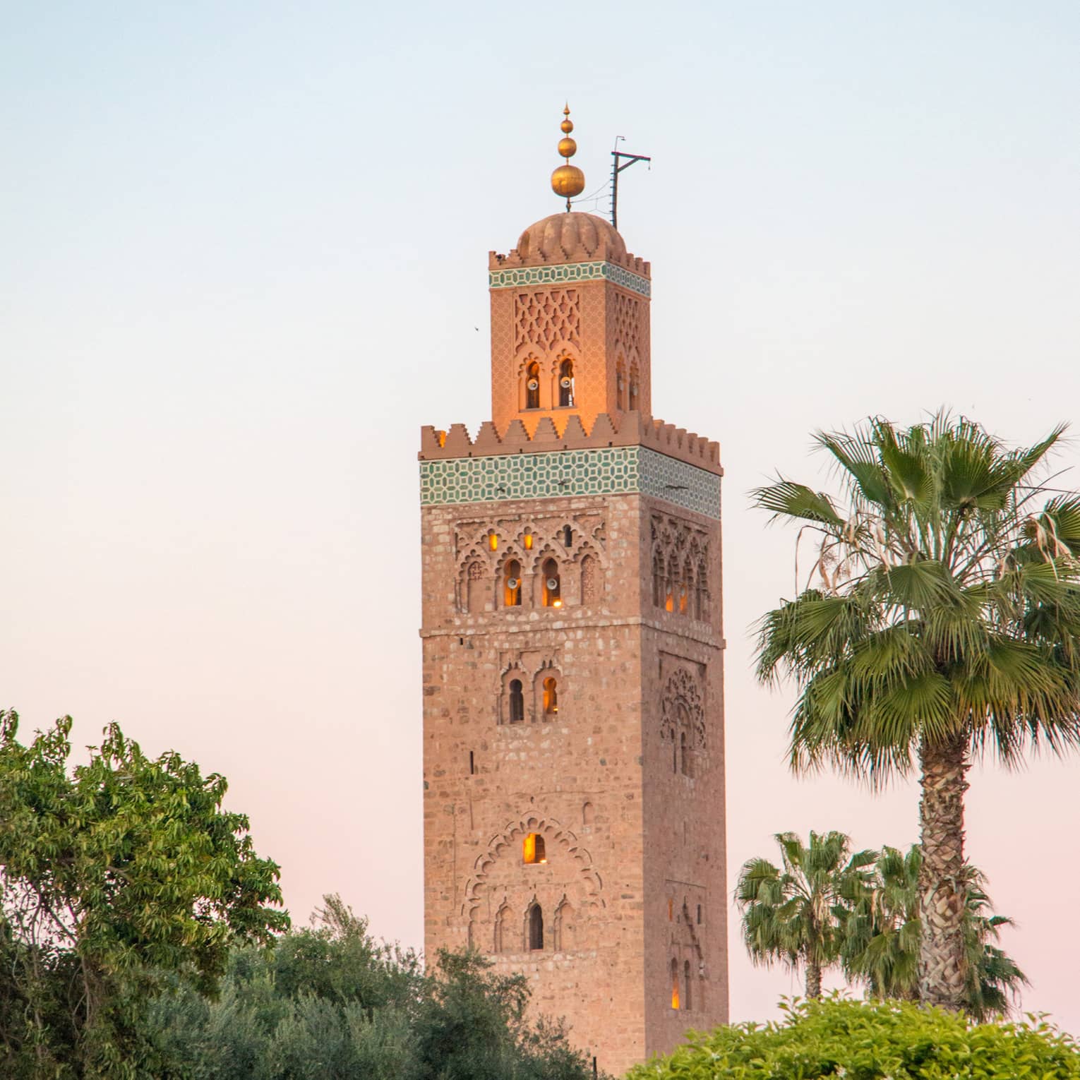 Rising from amid bushes and palms, a towering minaret with small domed archway windows, golden light emanating from within.