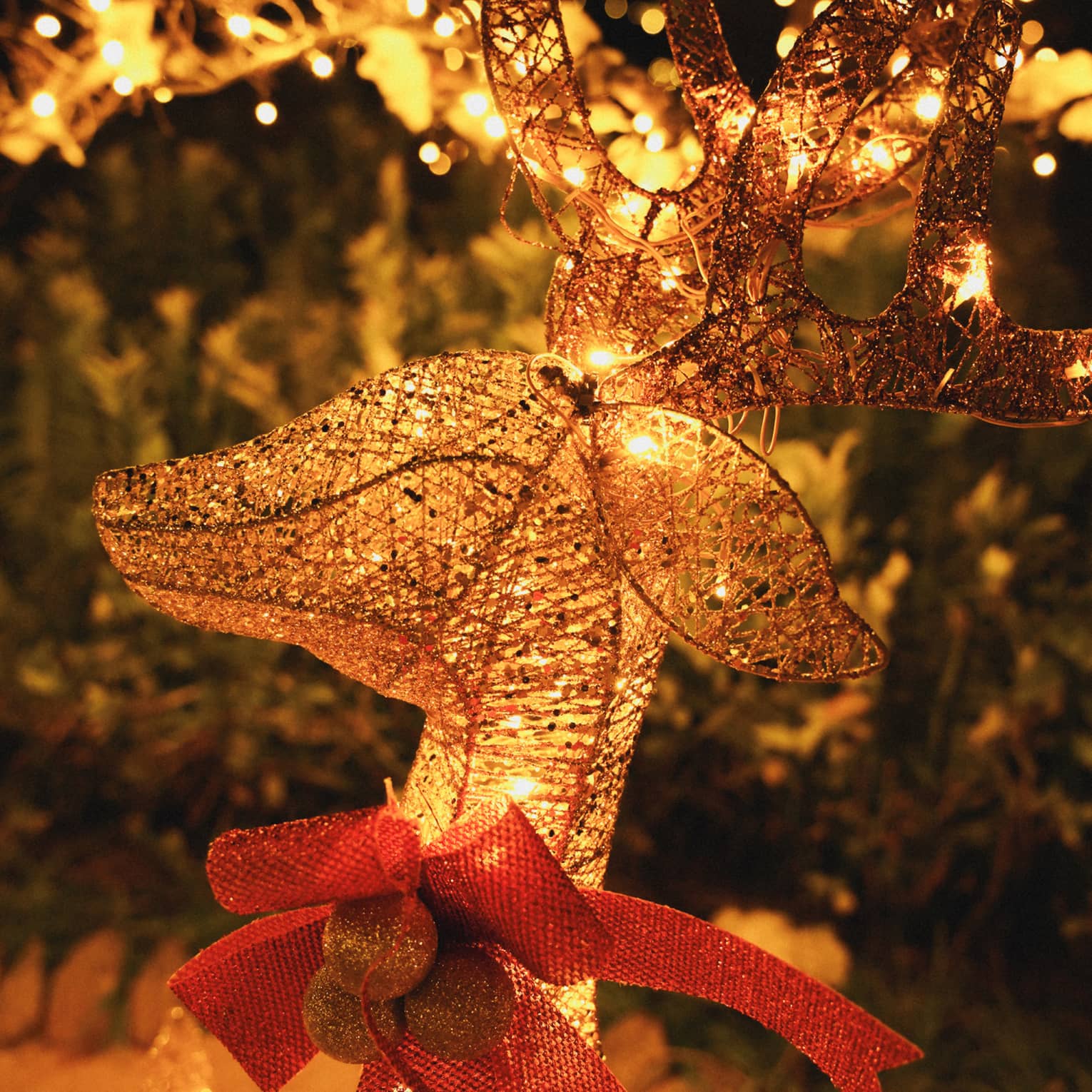 Close-up of a lit, golden wire reindeer wearing a large red bow with hazy festive greenery in the background.