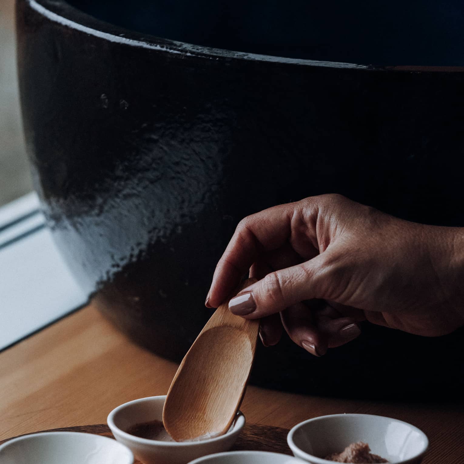 A hand places a wooden spoon into a small clay bowl to scoop up a spa ingredient, with three other clay bowls placed nearby