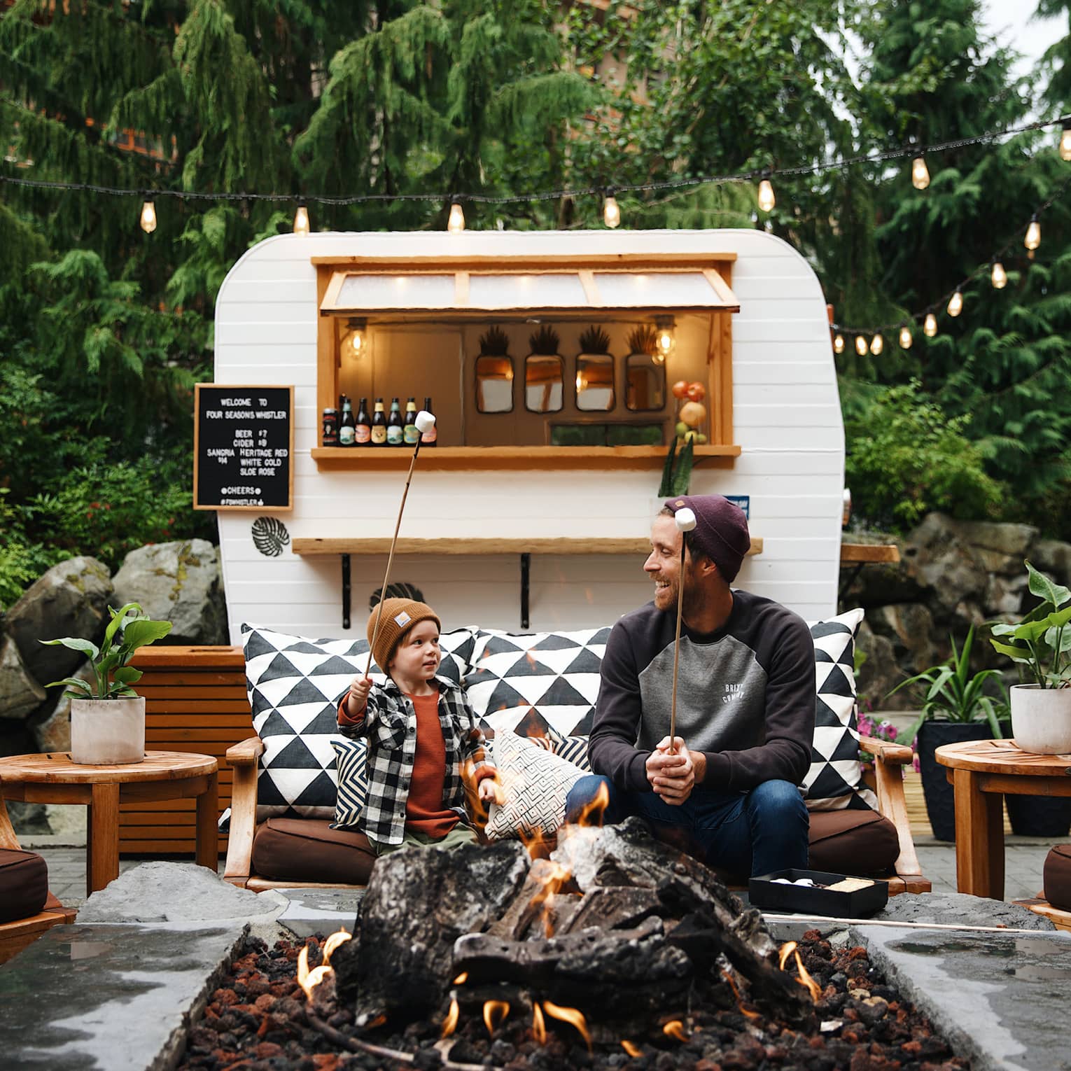 Adult and child sit on a bench by a fire in front of a small white camper serving s'mores