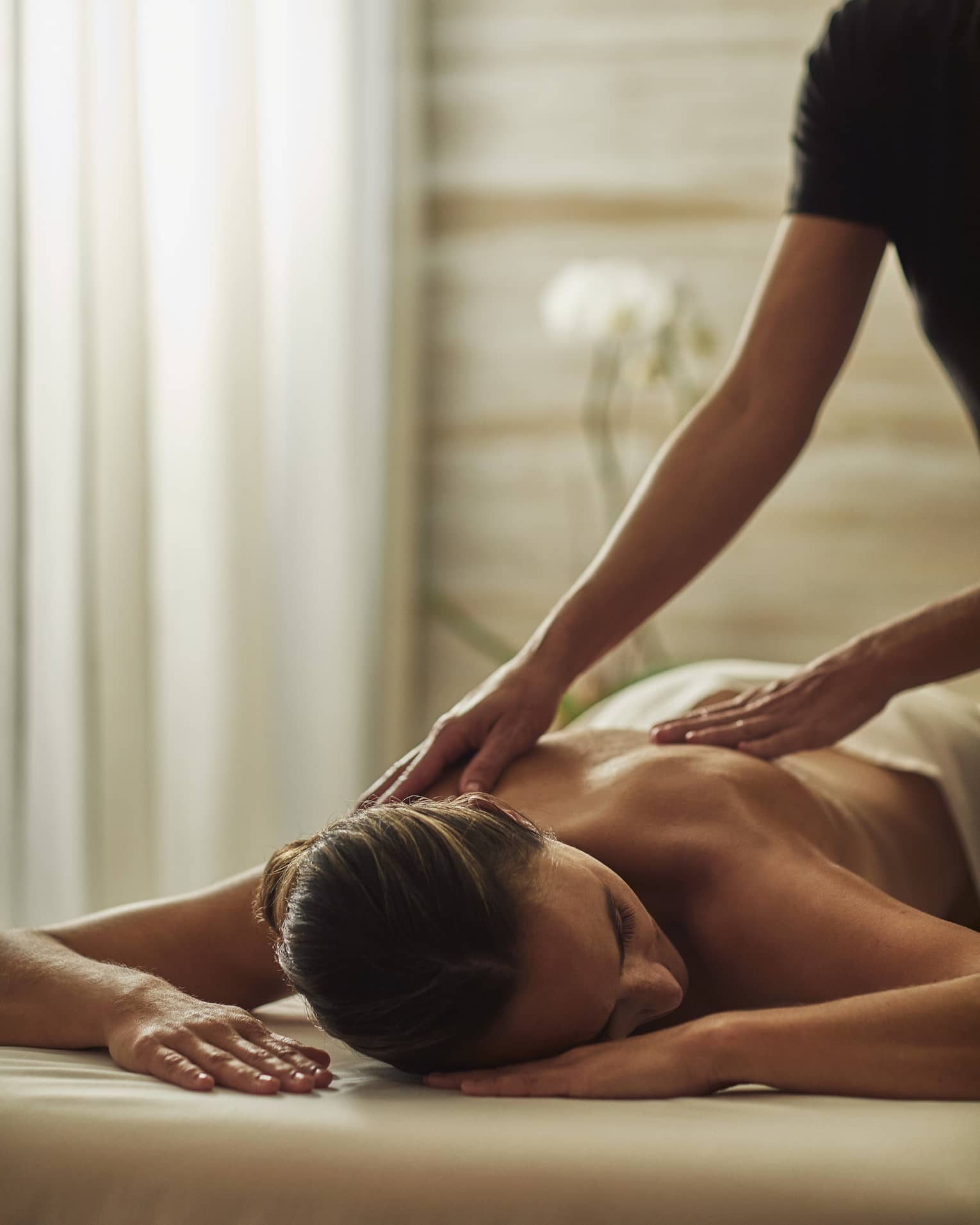 Woman in black uniform massages bare shoulders of woman lying on table face-down with head to side