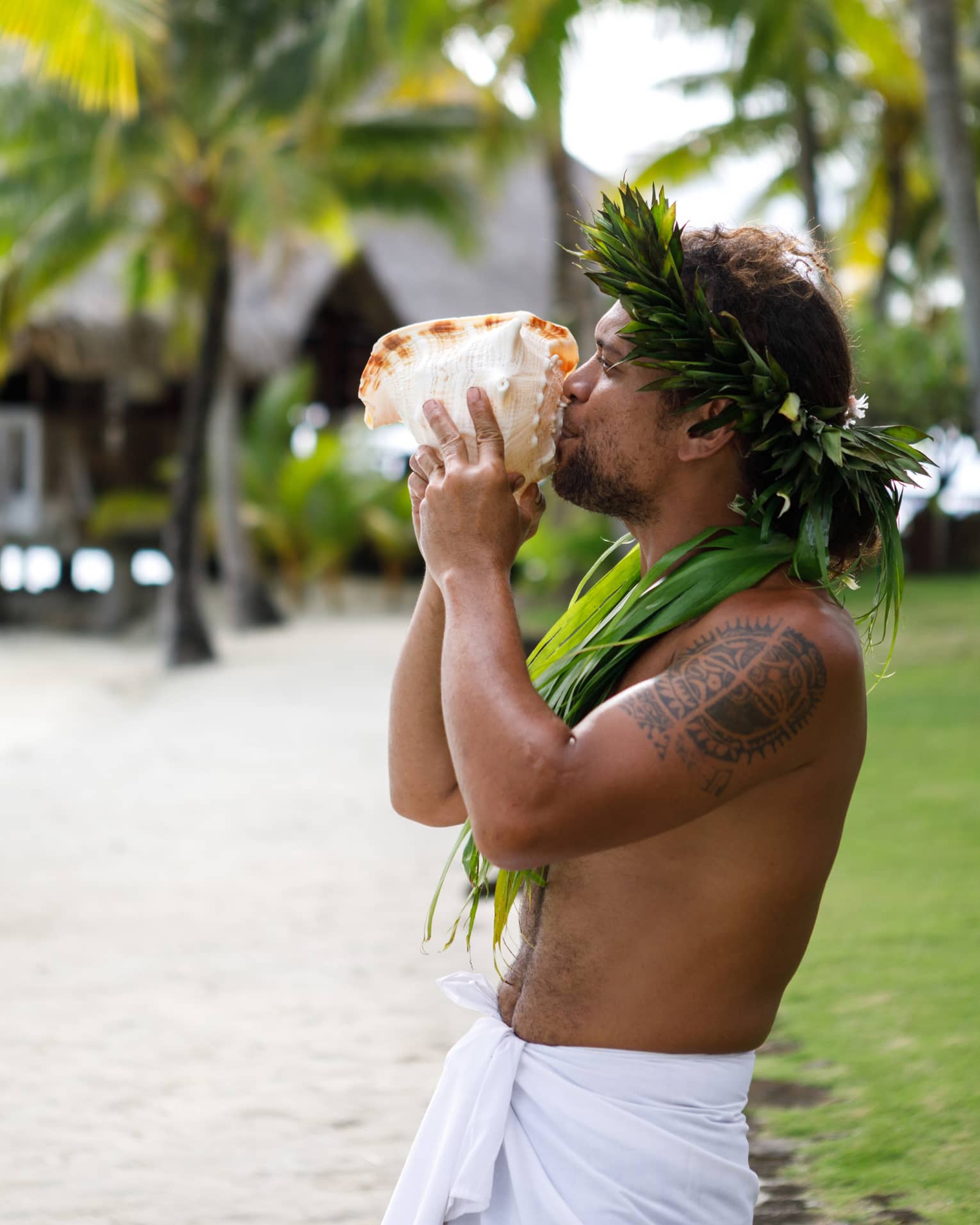 Tahitian man holds large sea shell to his lips on resort garden path