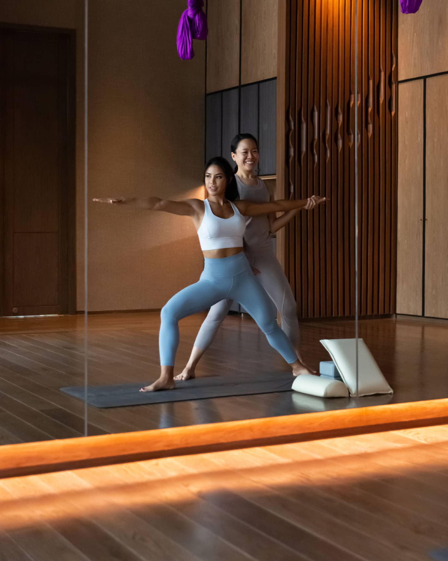 Mirror reflection of fitness staff helping woman stretch her arms in yoga pose under purple ribbons