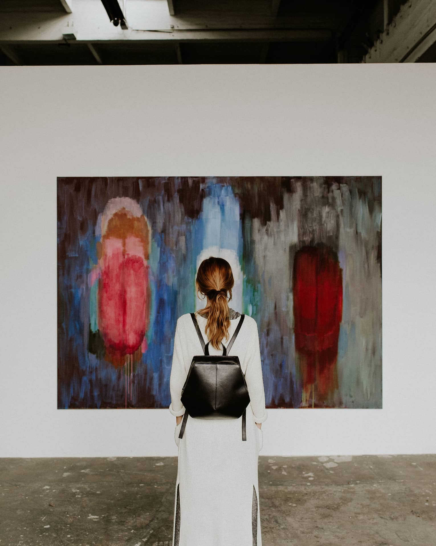 A person with a backpack standing in front of an abstract painting in an art gallery.
