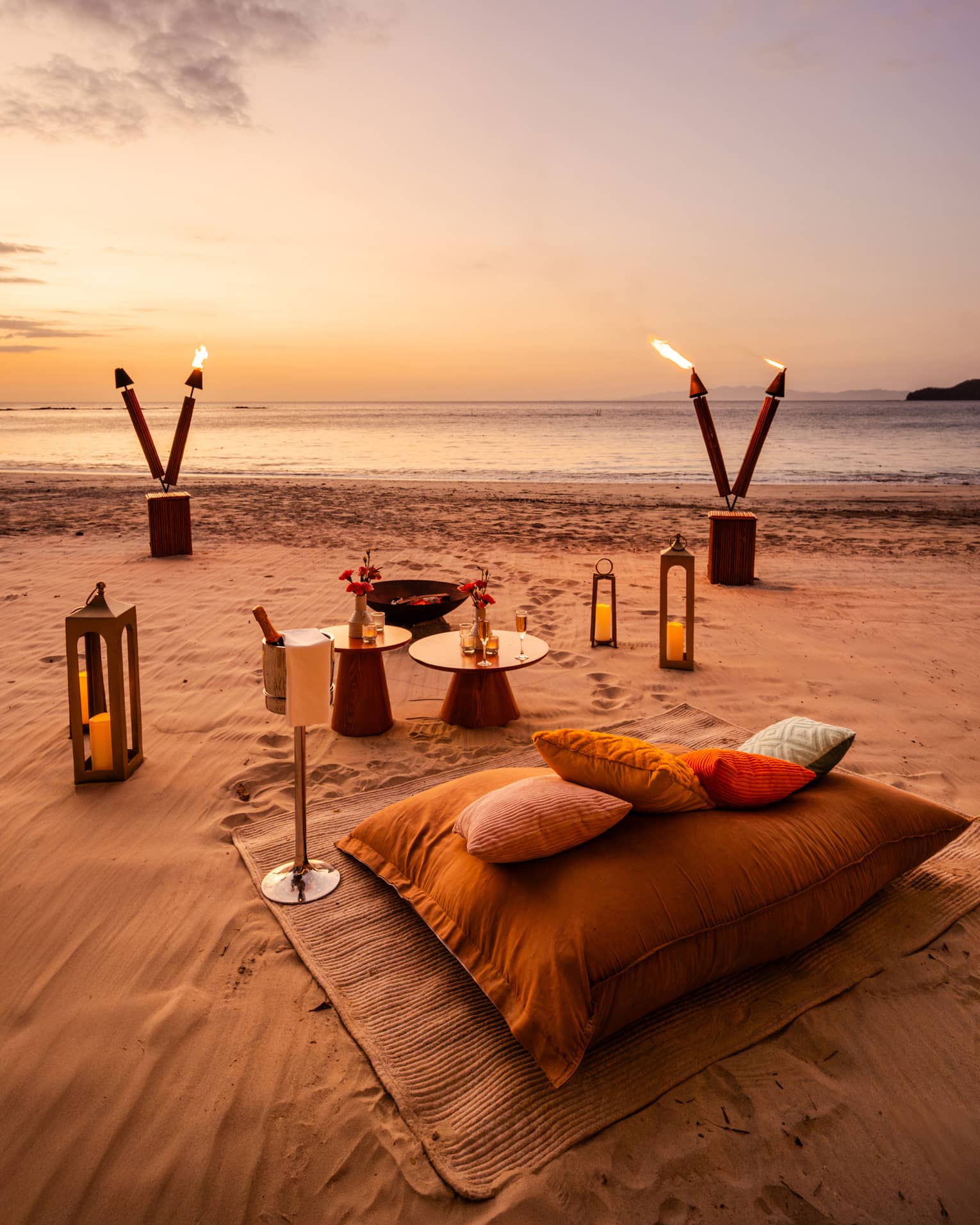 Blanket, cushions, small tables, lanterns and tiki torches are set up for a private dinner on a beach at sunset
