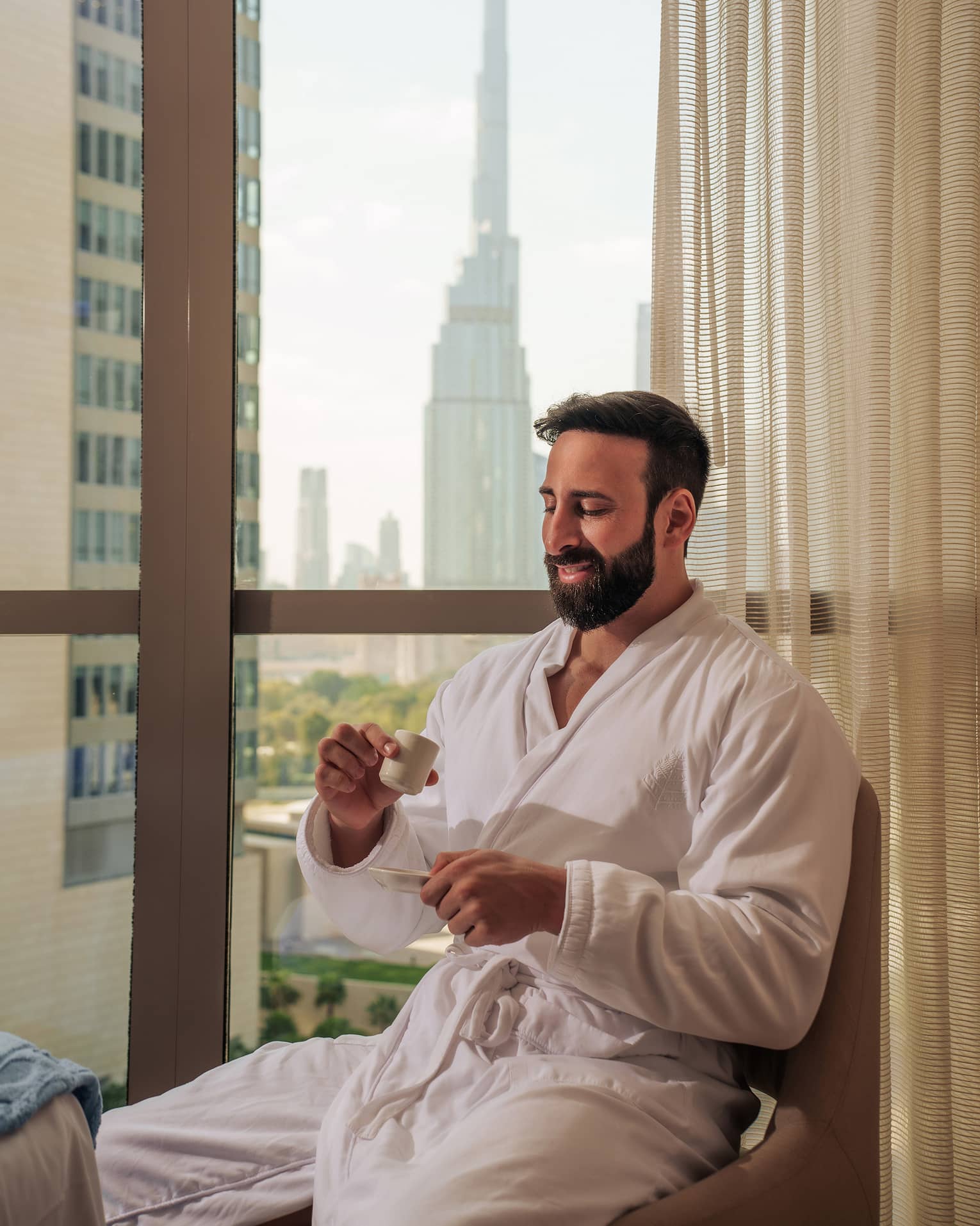 Person with short dark hair and beard and wearing a white spa bath robe sits by a window sipping tea with a city skyline in the background