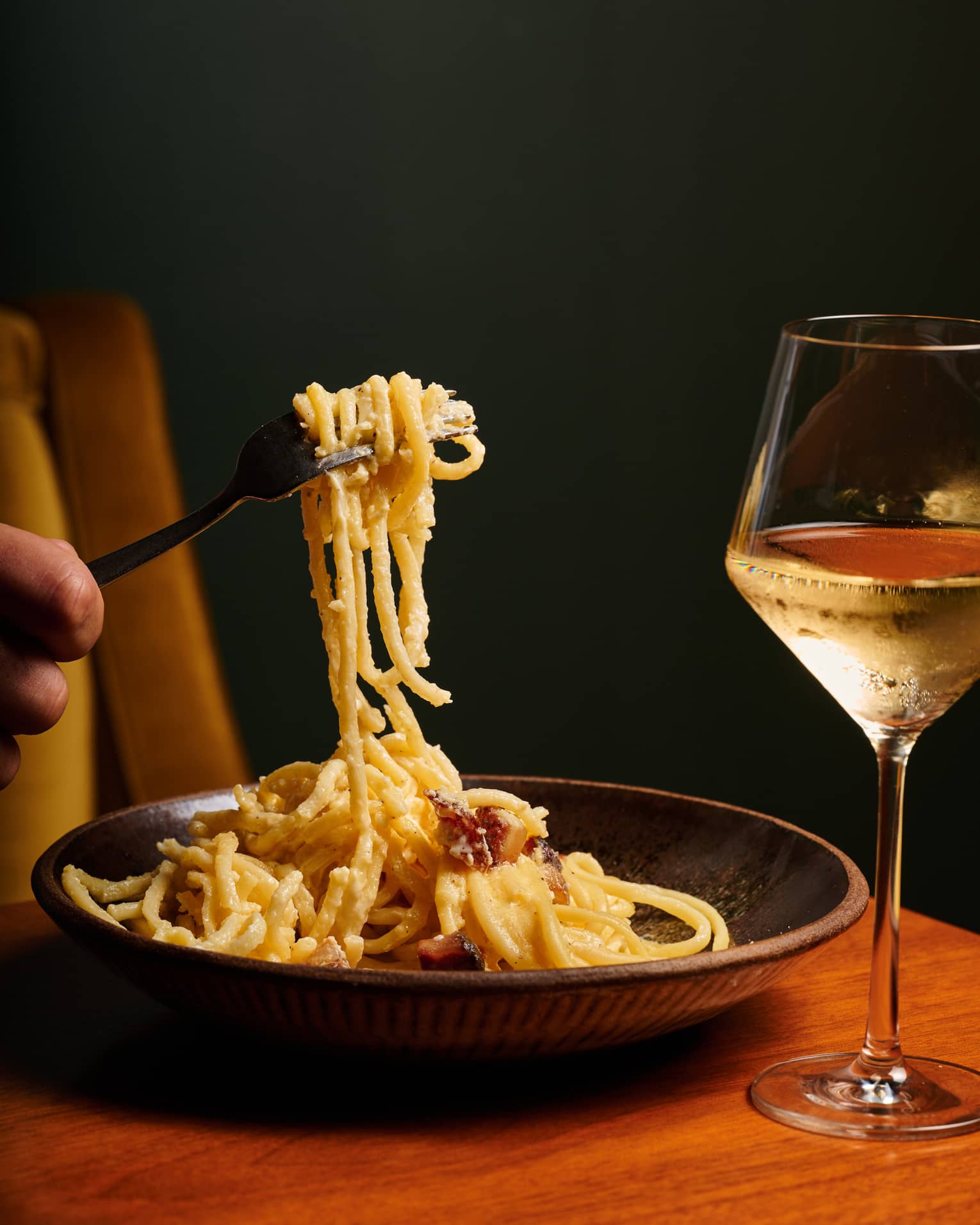 Close-up of a fork lifting spaghetti in a bowl of rich, creamy carbonara sauce with a glass of white wine to the right.