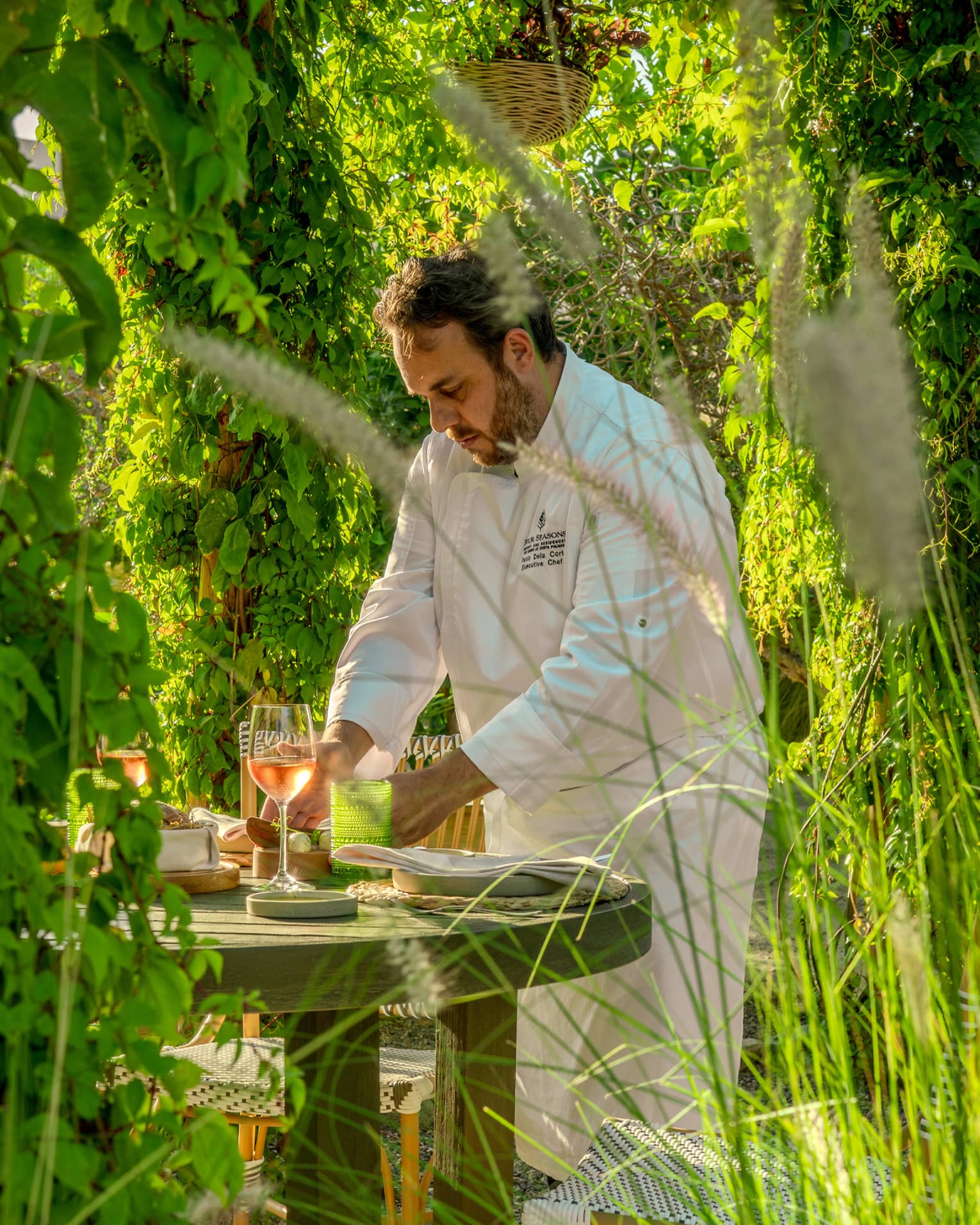 A chef works at a small dining table in a lush, green garden setting.