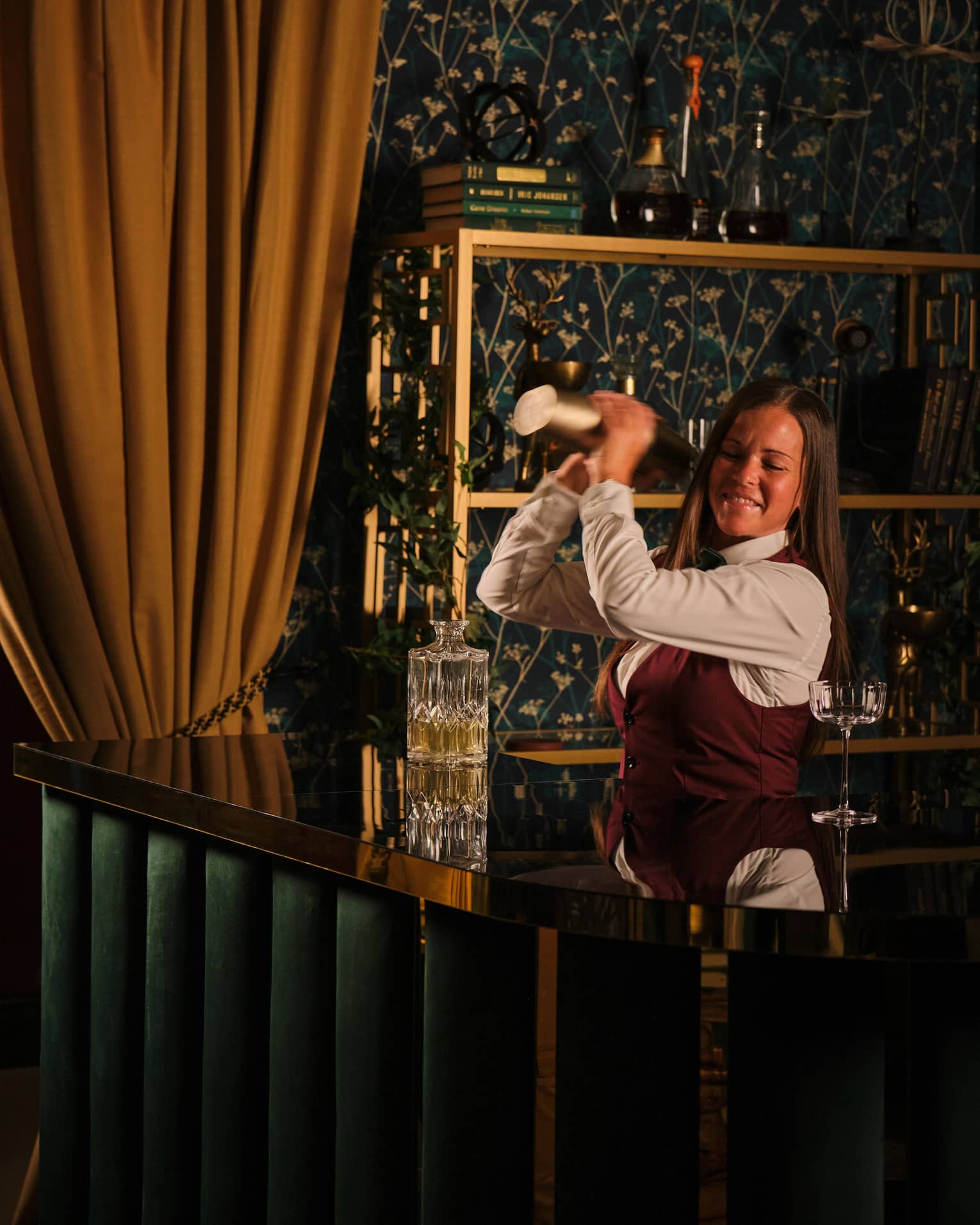 Bartender wearing white shirt, burgundy vest and black pants stand behind the bar shaking a cocktail