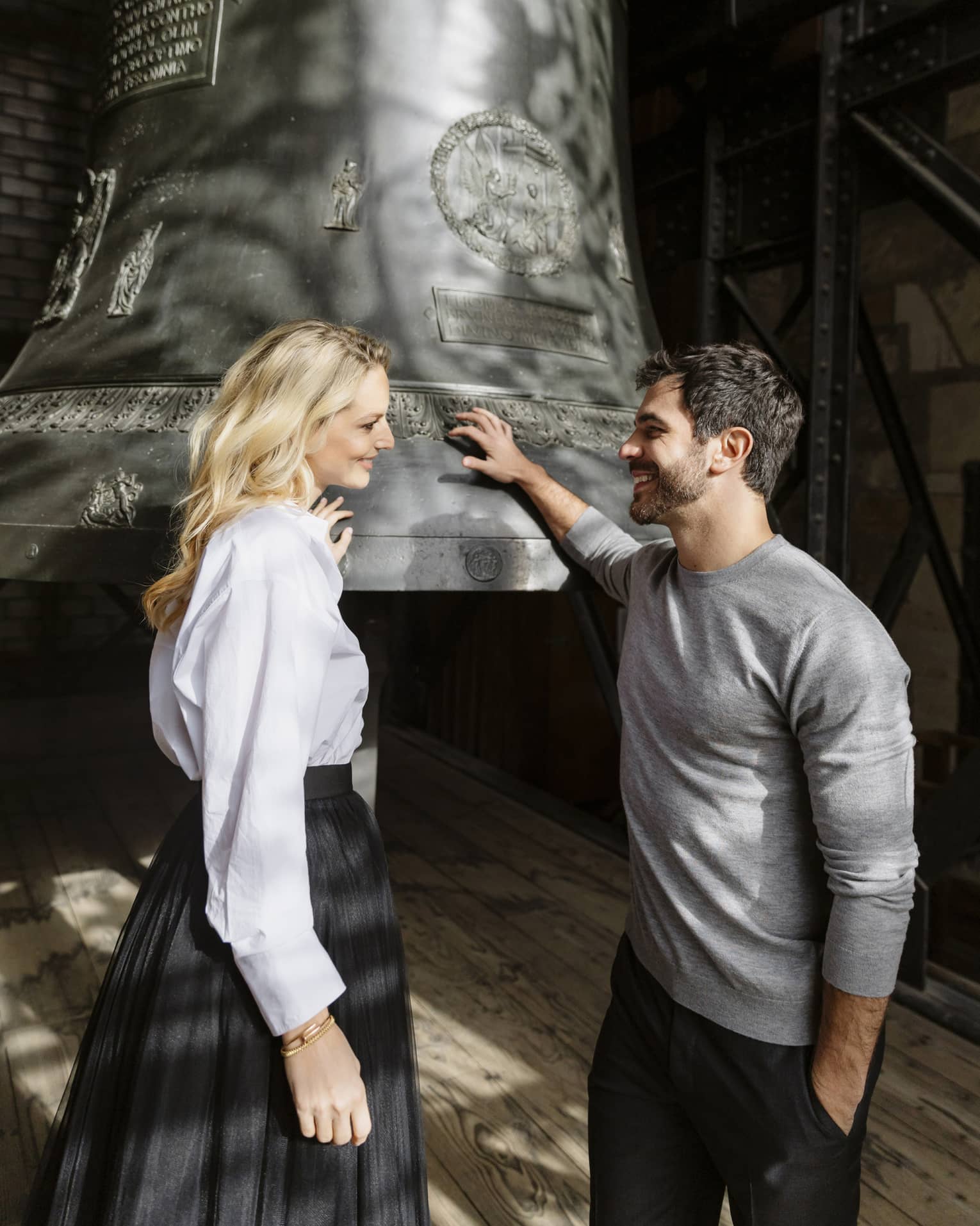 Two people stand next to a large metal bell in the bell tower of a cathedral.