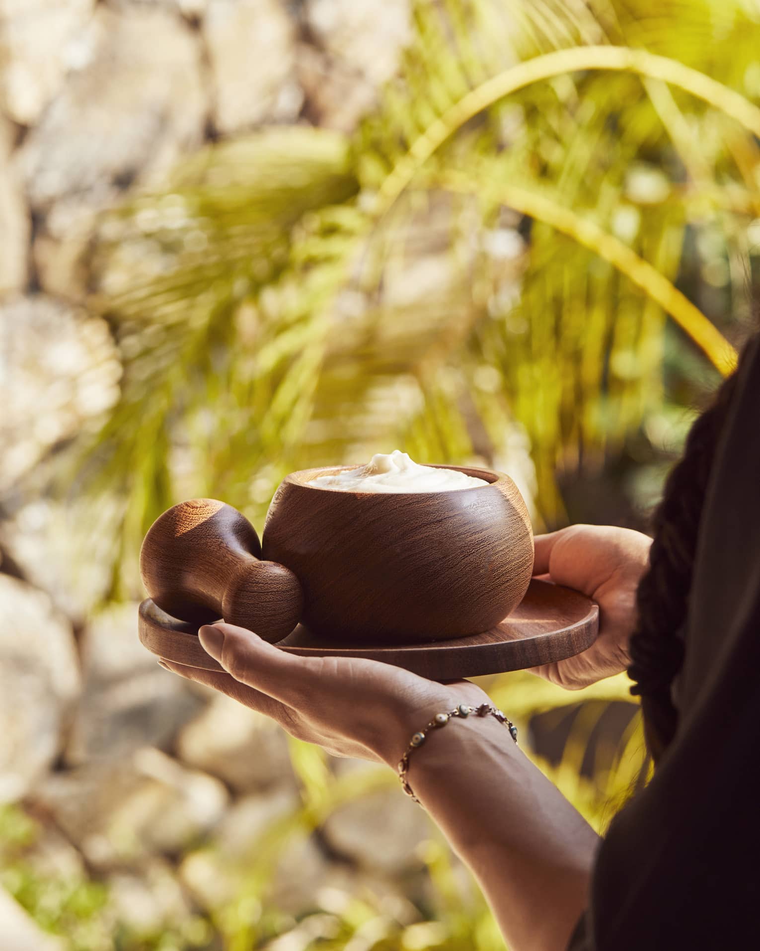 Two hands carry a small round tray with a wooden pestle and a bowl filled with a creamy body scrub past golden palm fronds.
