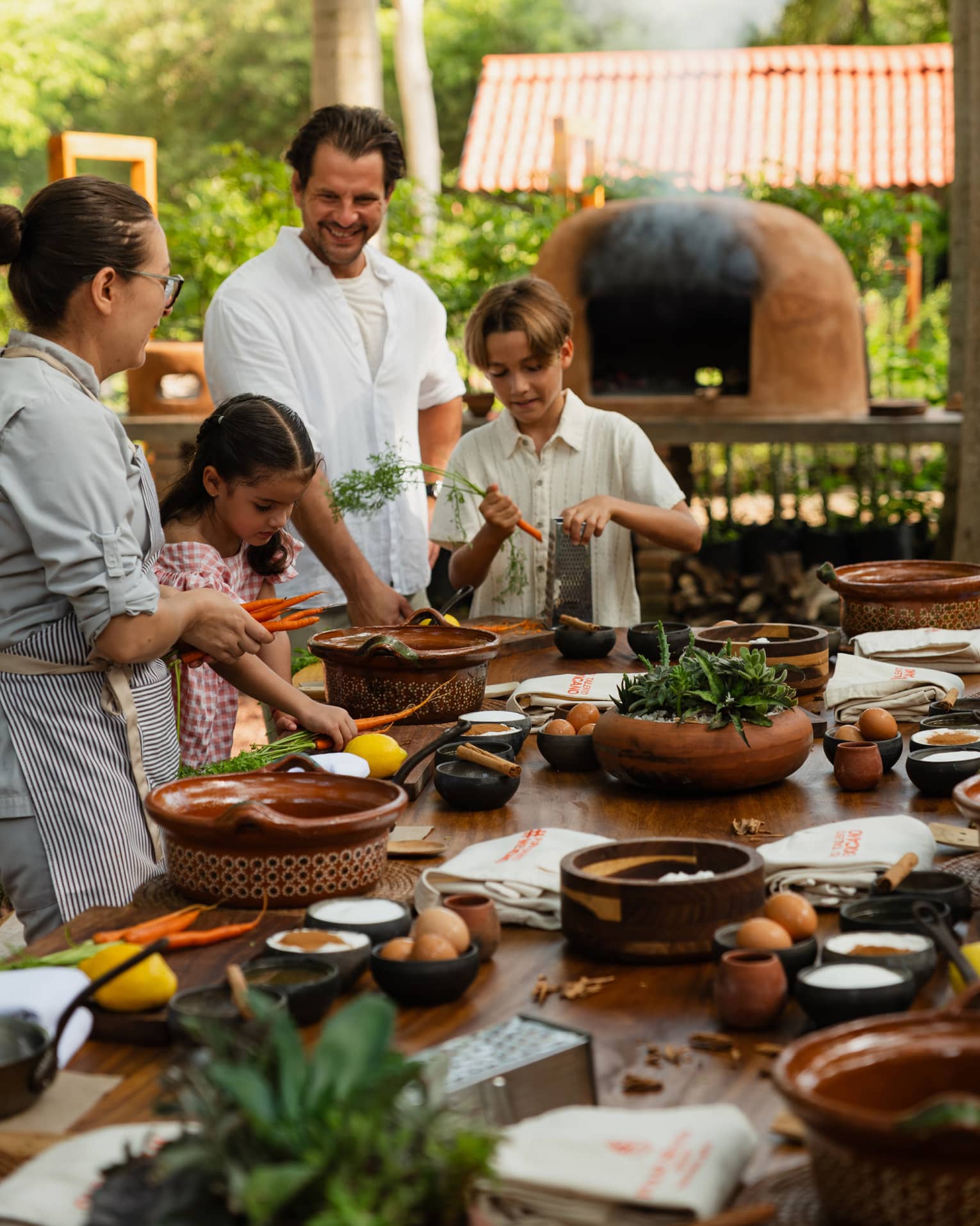 Adults and children standing around an outdoor dining table with various dishes and utensils on it, while an outdoor oven runs in the background.